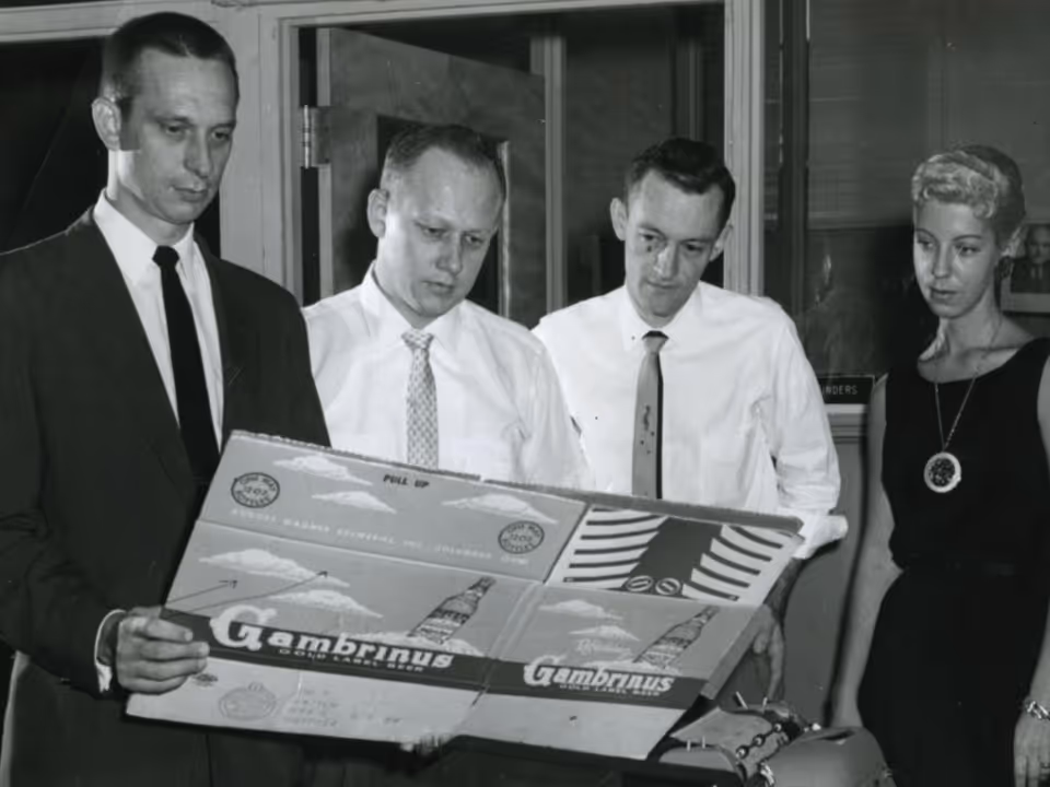 Four people in 1960s attire closely examining a large cardboard package labeled 'Gambrinus Gold Label Beer'.