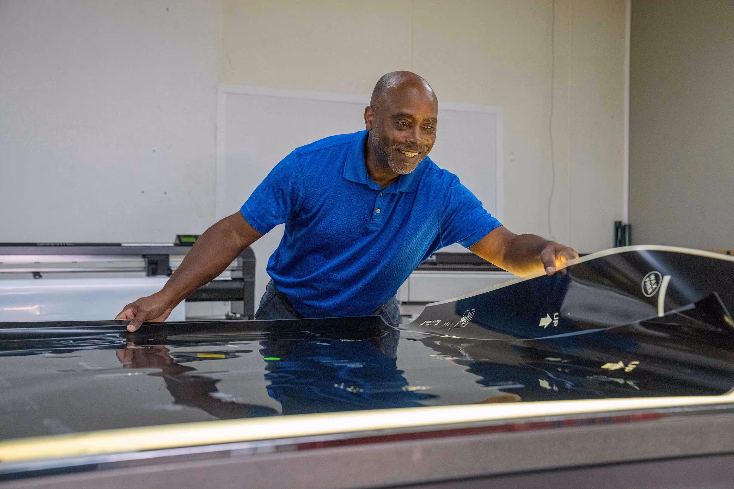Man in blue polo shirt carefully peeling back a large black film or sheet in a workspace.