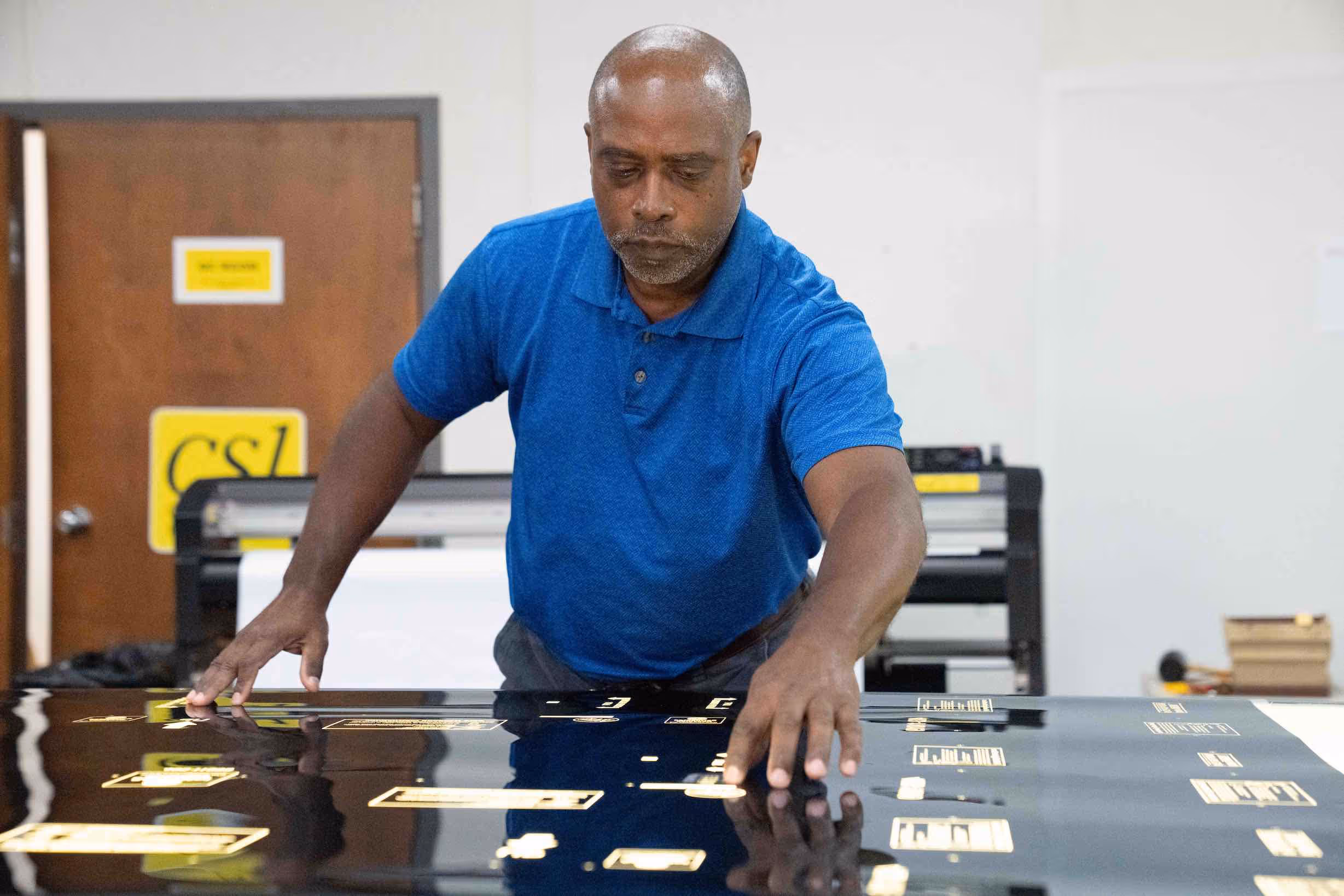 Man in blue polo shirt positioning a large black glossy sheet with gold printed labels on a table.