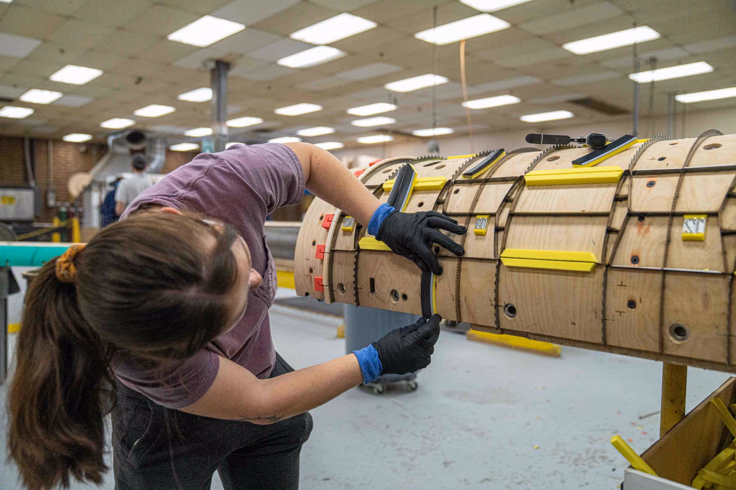 Person wearing black gloves assembling or inspecting a curved wooden structure with yellow and black components in a workshop.
