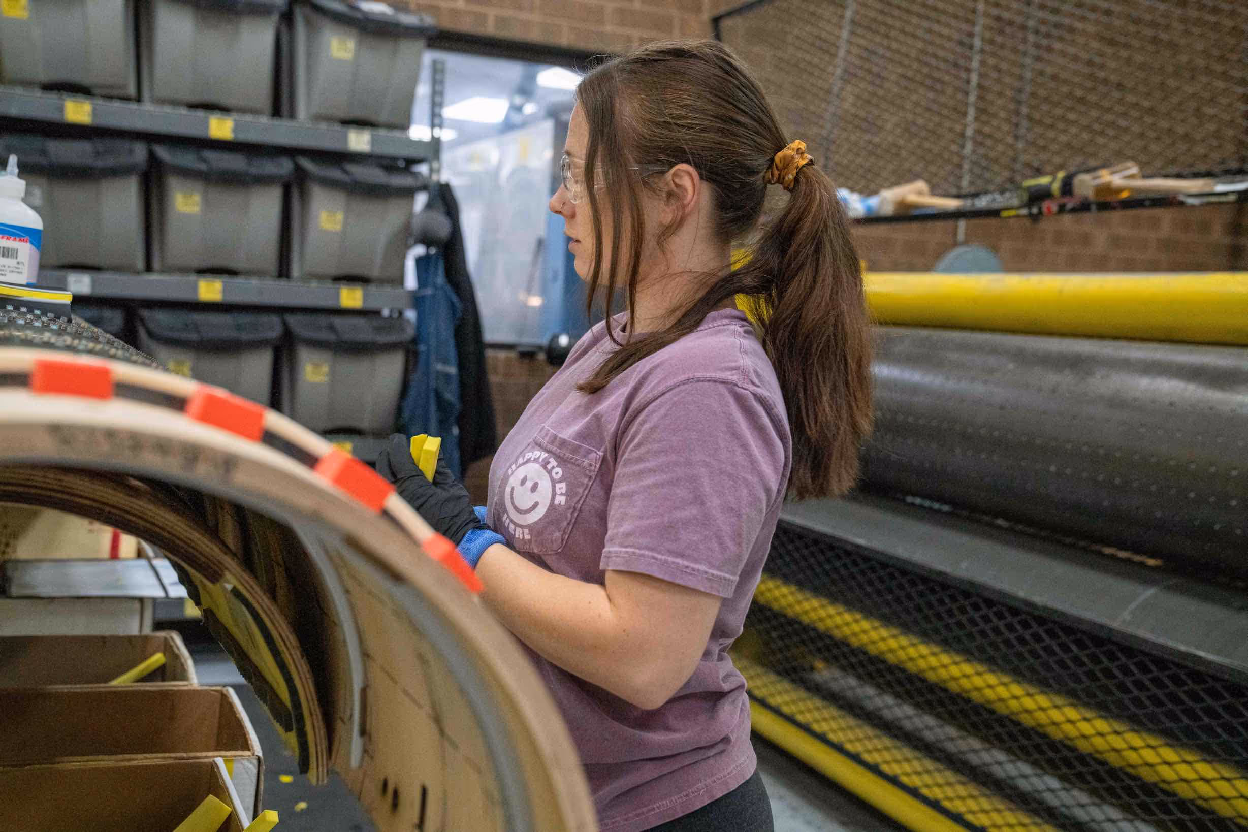 Woman wearing safety glasses and gloves working with curved wooden pieces in a workshop.