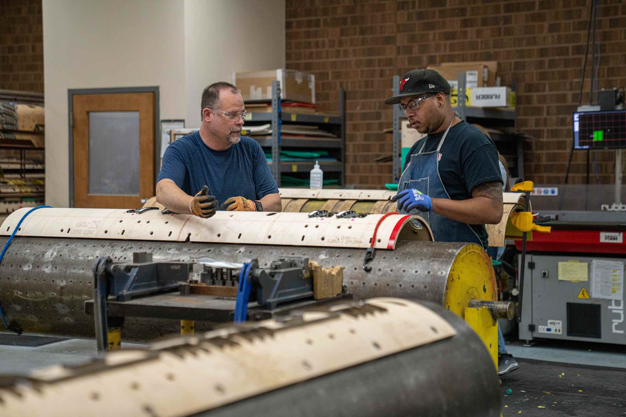 Two factory workers examining and discussing a large cylindrical metal part in an industrial workshop.
