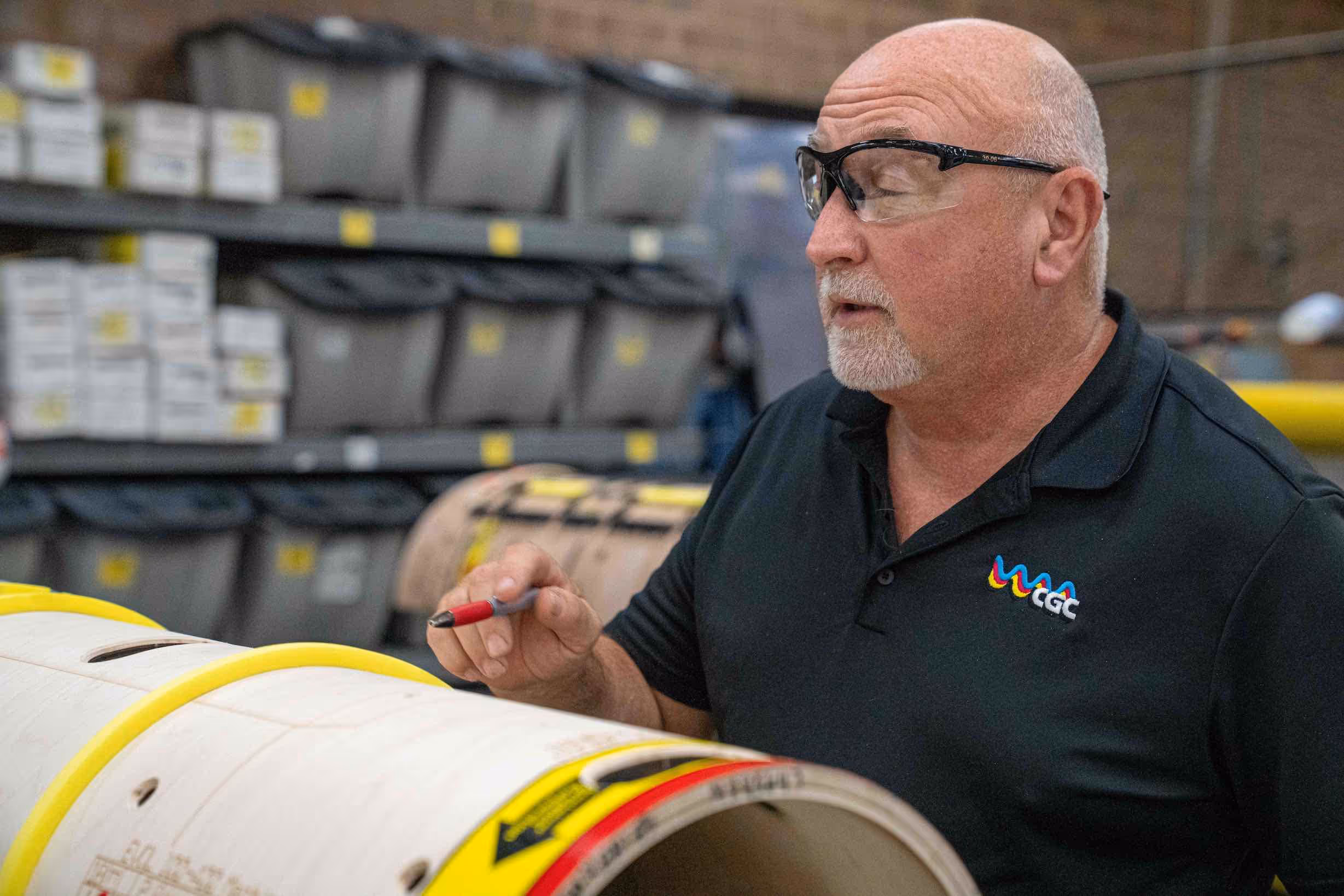 Man wearing safety glasses and a black GGC polo shirt inspecting a large cylindrical object with yellow edges in a storage area.