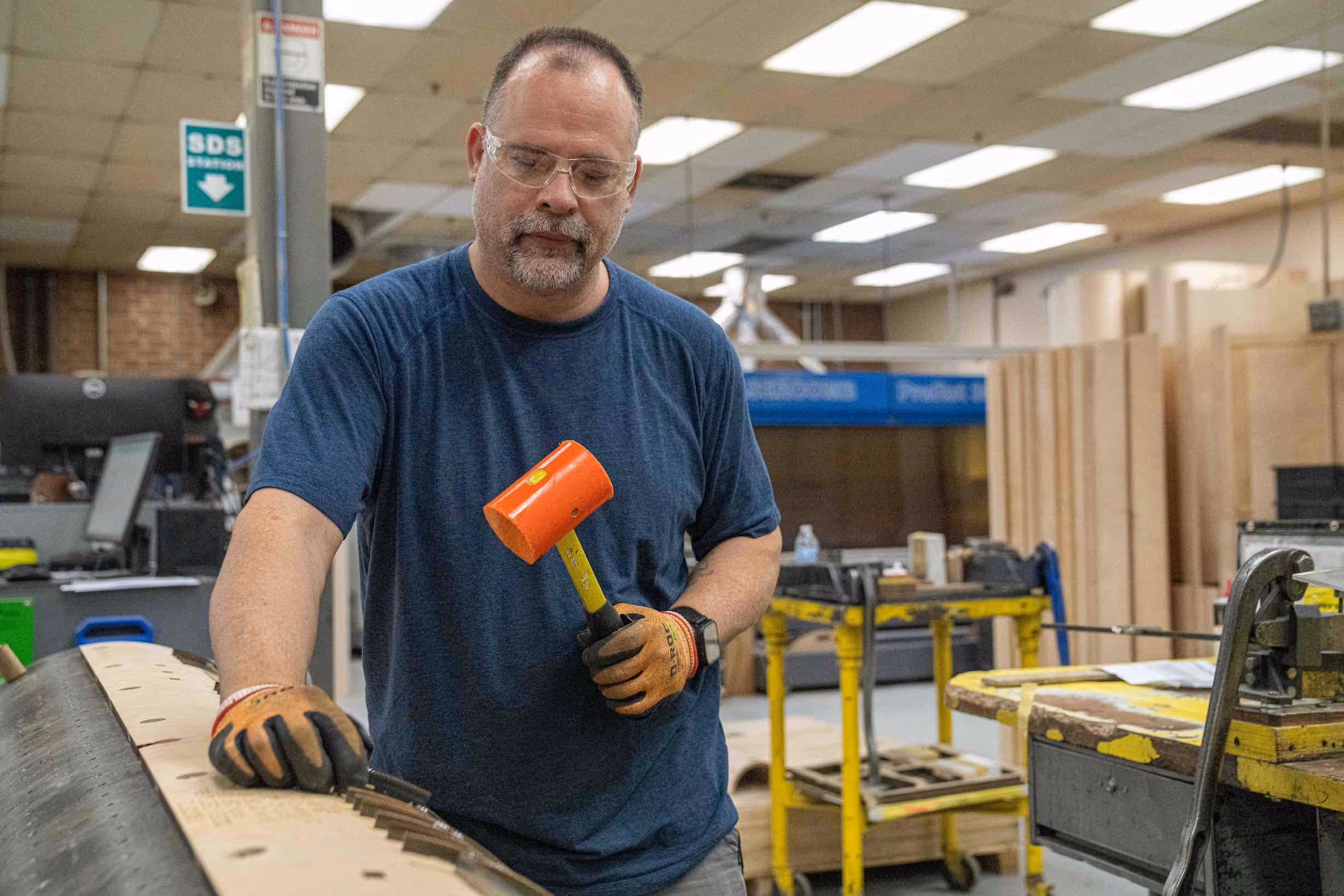 Man wearing safety glasses and gloves holds an orange mallet while working on a large curved wooden piece in a workshop.