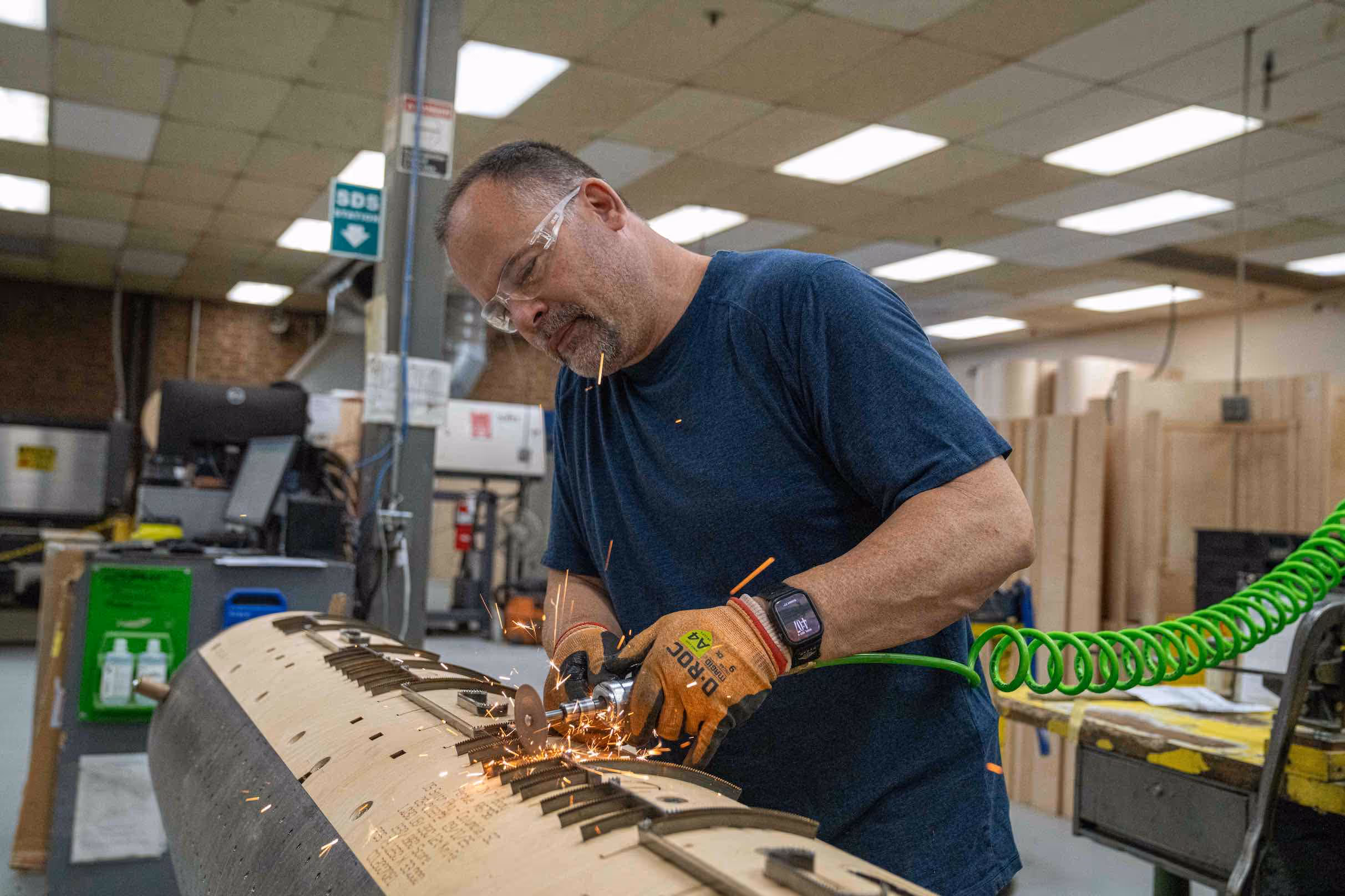Man wearing safety glasses and gloves using a handheld grinder producing sparks on a curved wooden surface in a workshop.