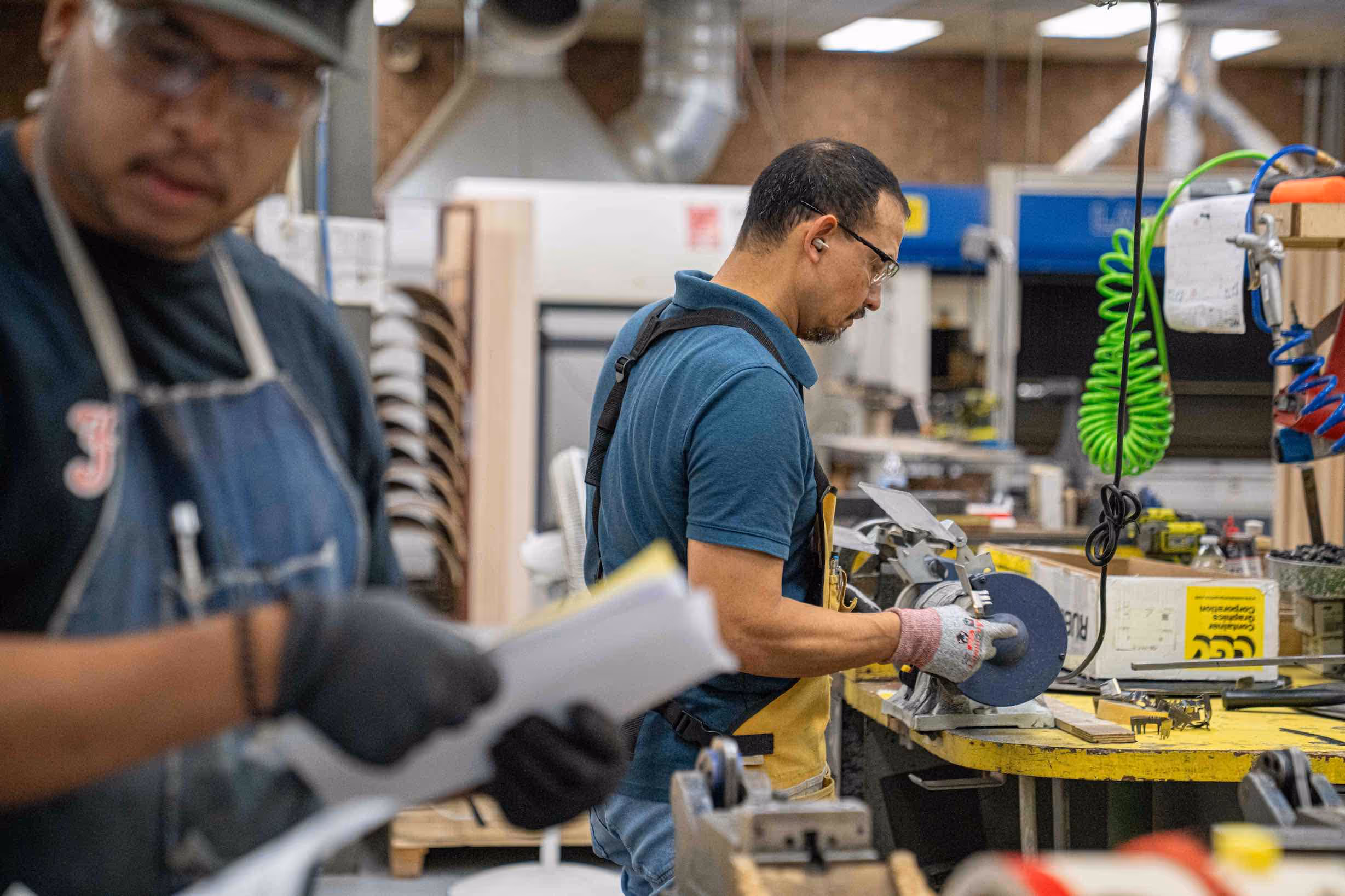 Two factory workers in safety glasses and gloves operating machinery and handling documents in a workshop.
