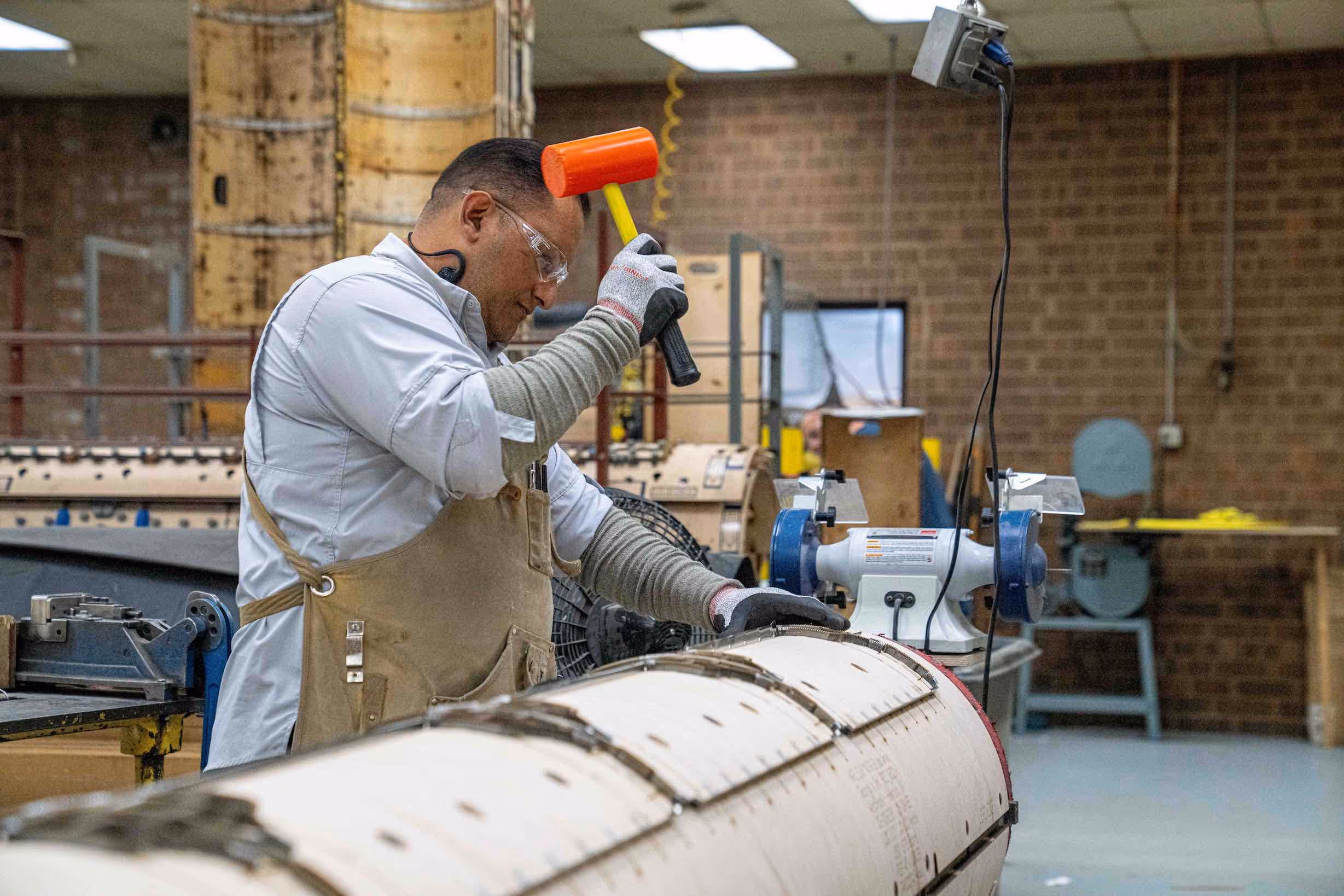 Worker wearing safety glasses and gloves uses an orange mallet on a large cylindrical mold in a workshop.