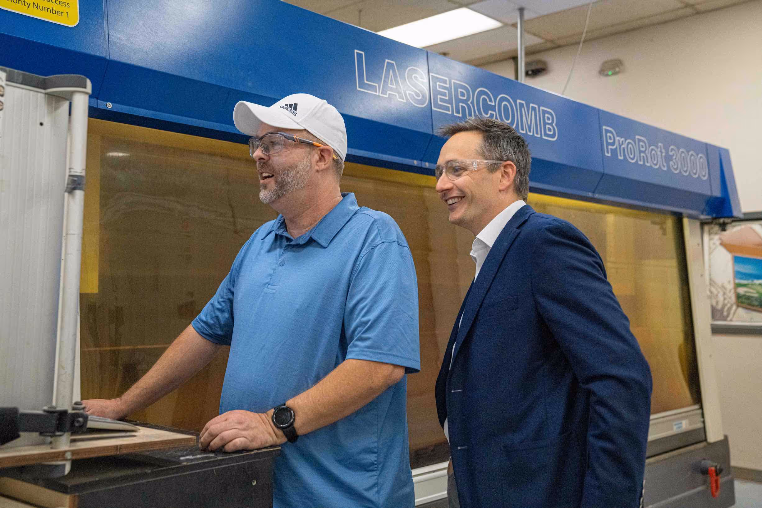 Two men wearing safety glasses stand by a LASERCOM ProRot 3000 industrial machine, engaged in conversation and smiling.