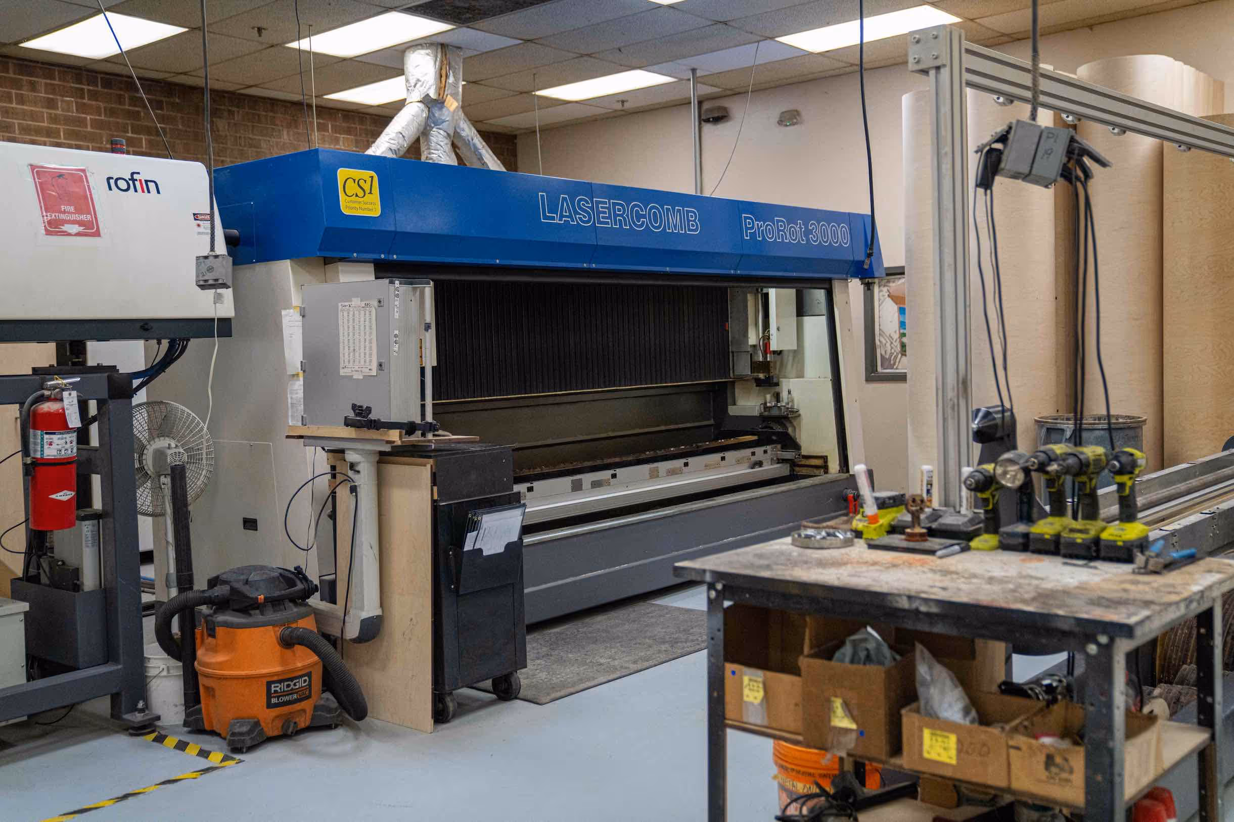 Industrial workshop featuring a large LASERCOMB ProRot 3000 laser cutting machine and a workbench with multiple power drills and tools.