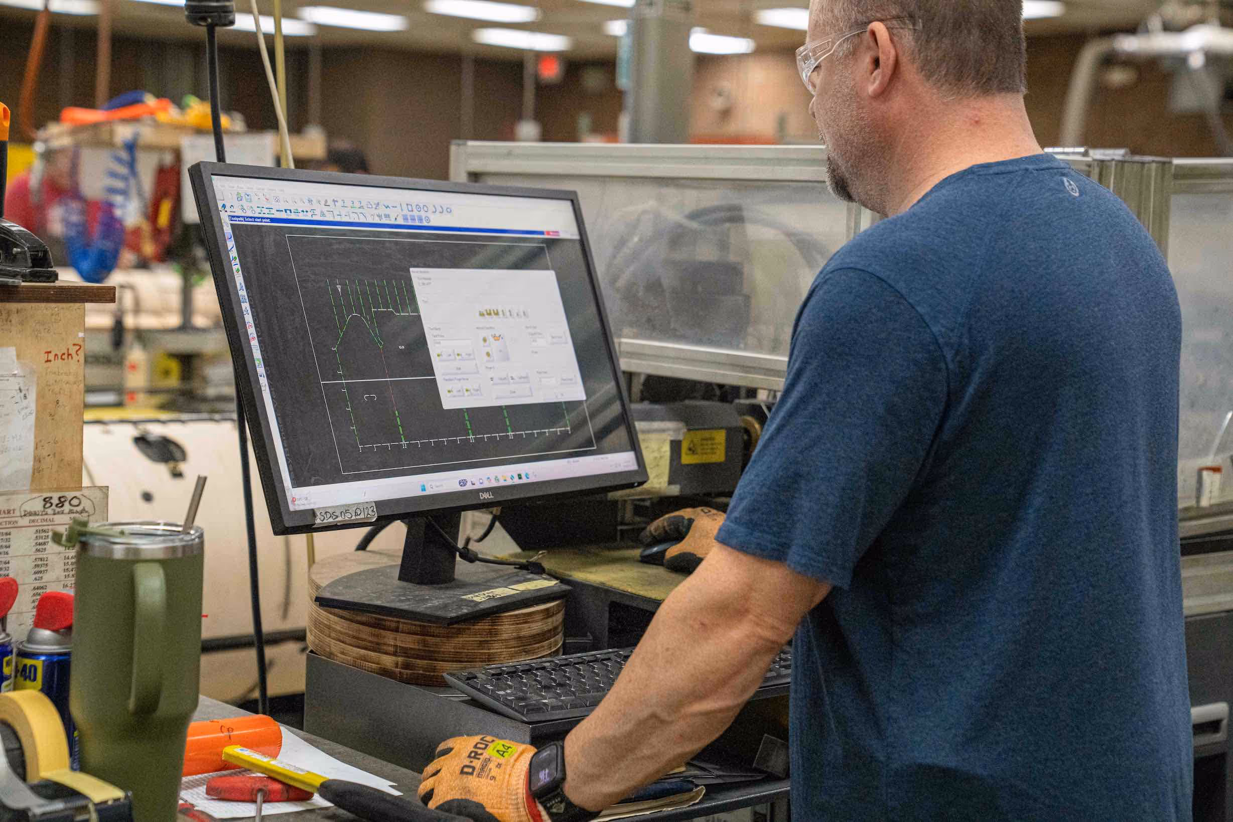 Man wearing safety glasses and gloves working on a computer with technical design software in an industrial workshop.