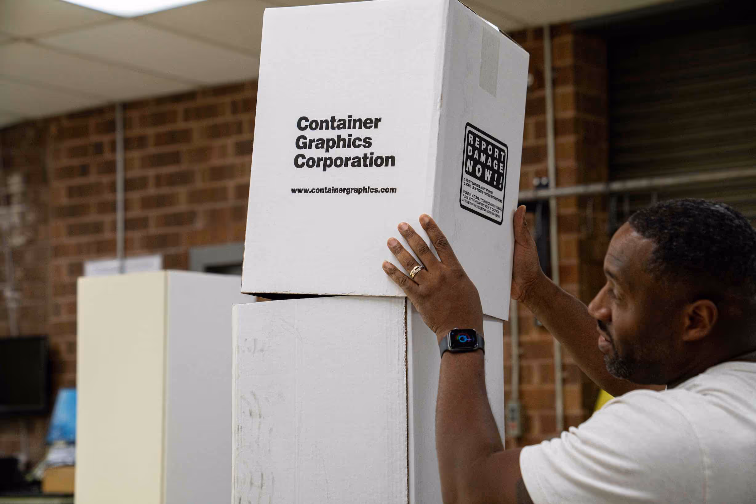 Man stacking white cardboard boxes labeled Container Graphics Corporation in a warehouse with brick walls.