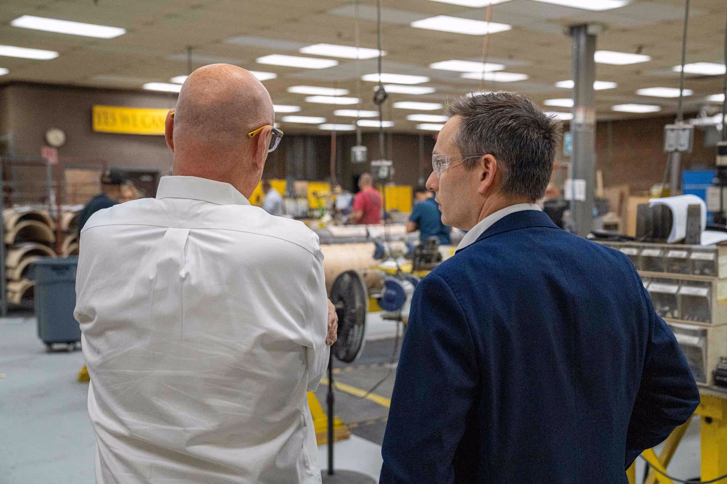 Two men wearing safety glasses discussing in a manufacturing facility with workers and machinery in the background.