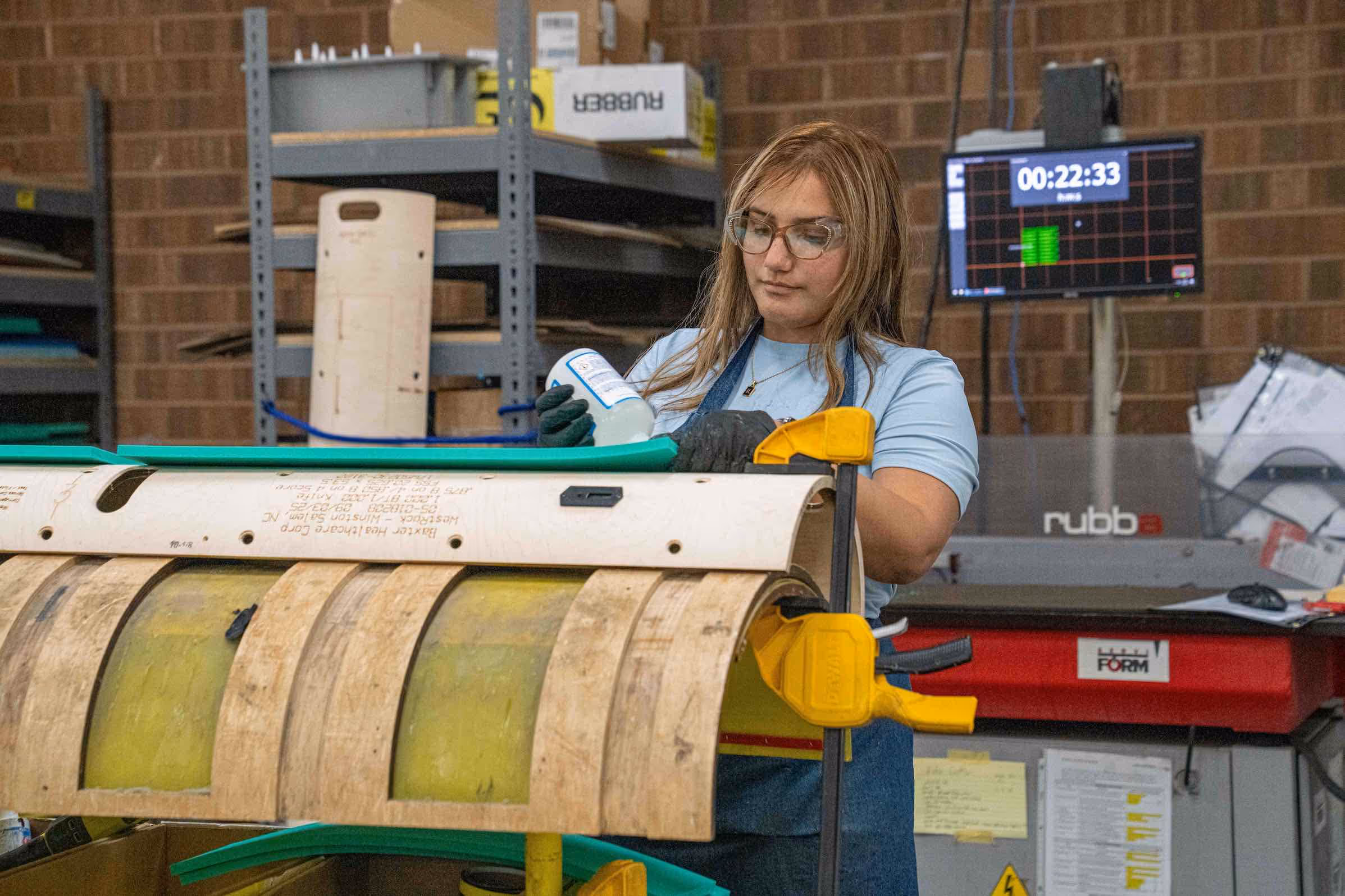 Woman in safety glasses and gloves working with a curved mold and green material in an industrial workshop.