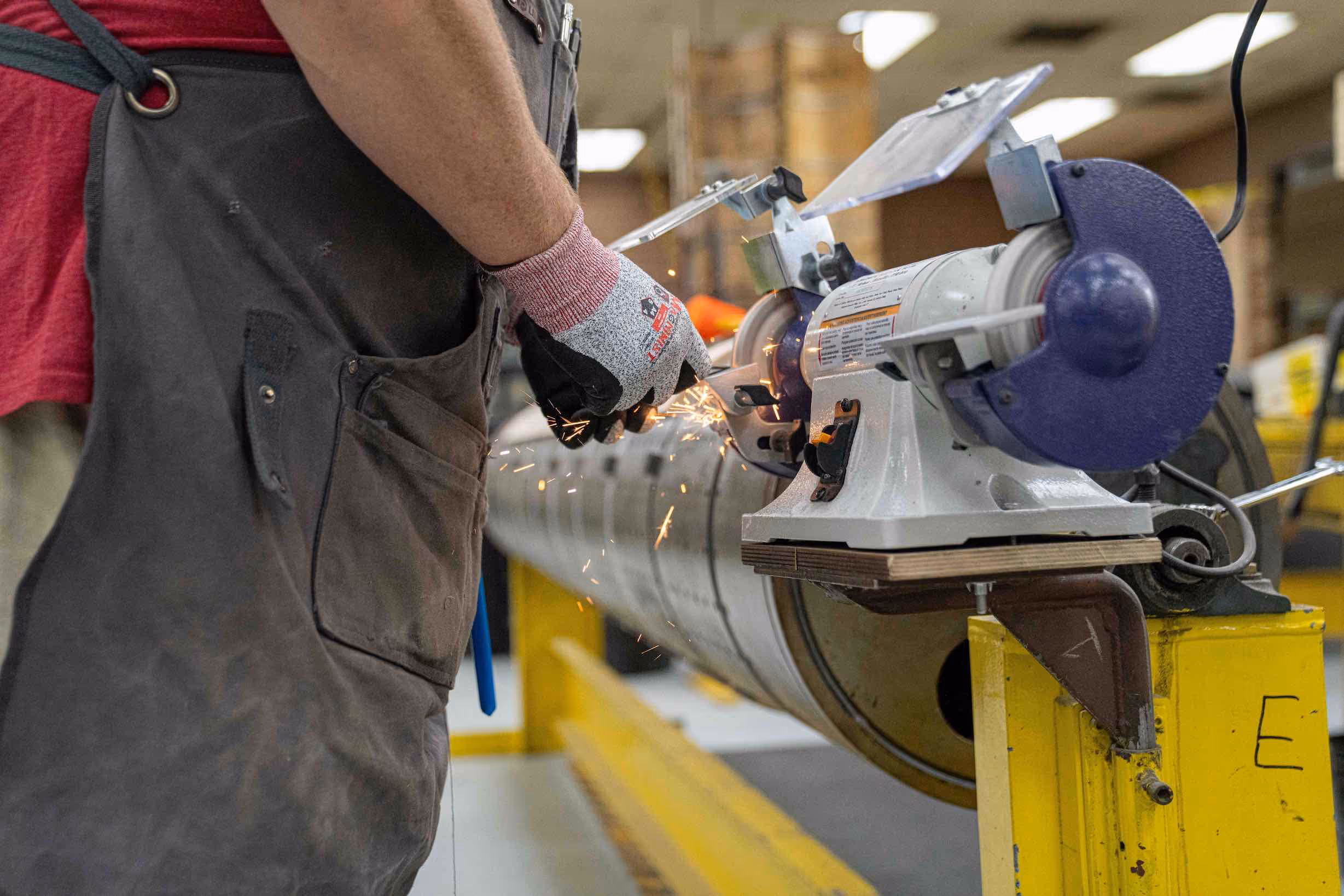 Worker wearing protective gloves using a bench grinder to sharpen or grind a metal object, with sparks flying.