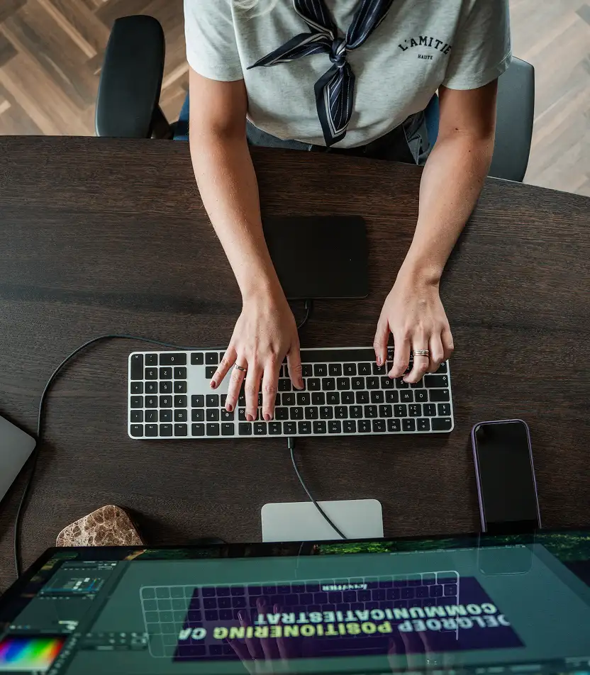 Bovenaanzicht van een persoon die op een toetsenbord typt achter een computerscherm op een houten bureau.