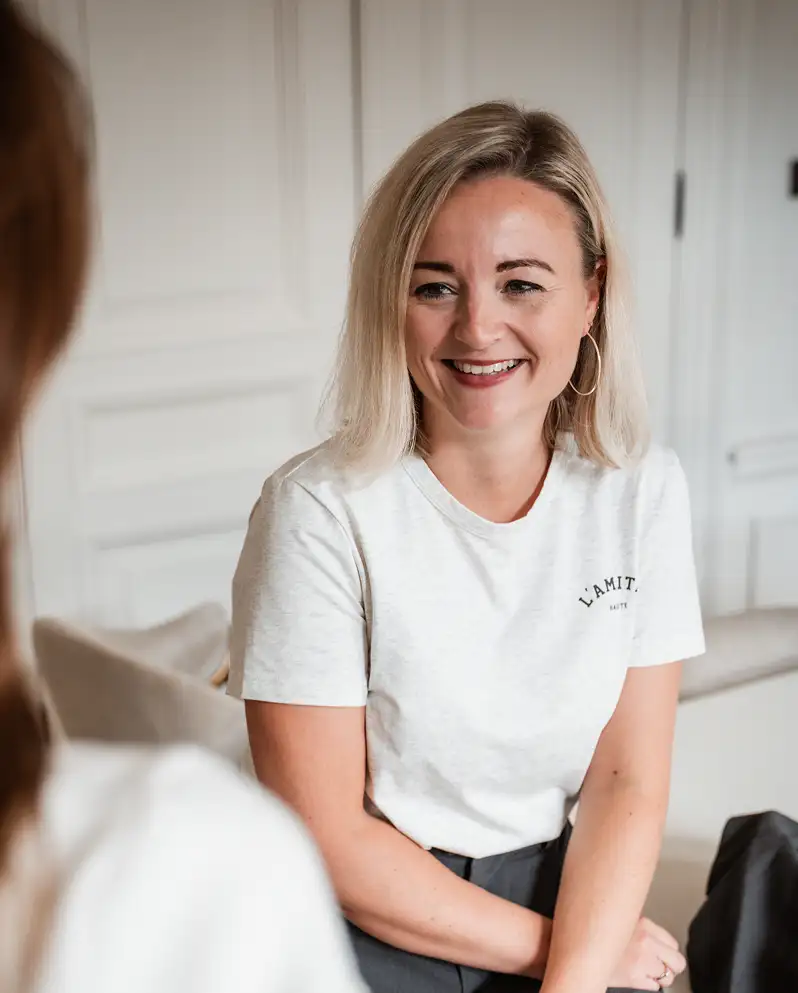 Vrouw met blond haar en witte T-shirt lachend tijdens gesprek in een lichte kamer.