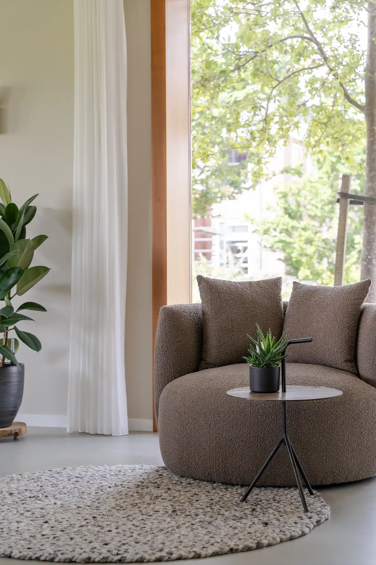 Comfortabele ronde bruine stoel met kussens, klein houten tafeltje met plant en grote raam met uitzicht op groene bomen.
