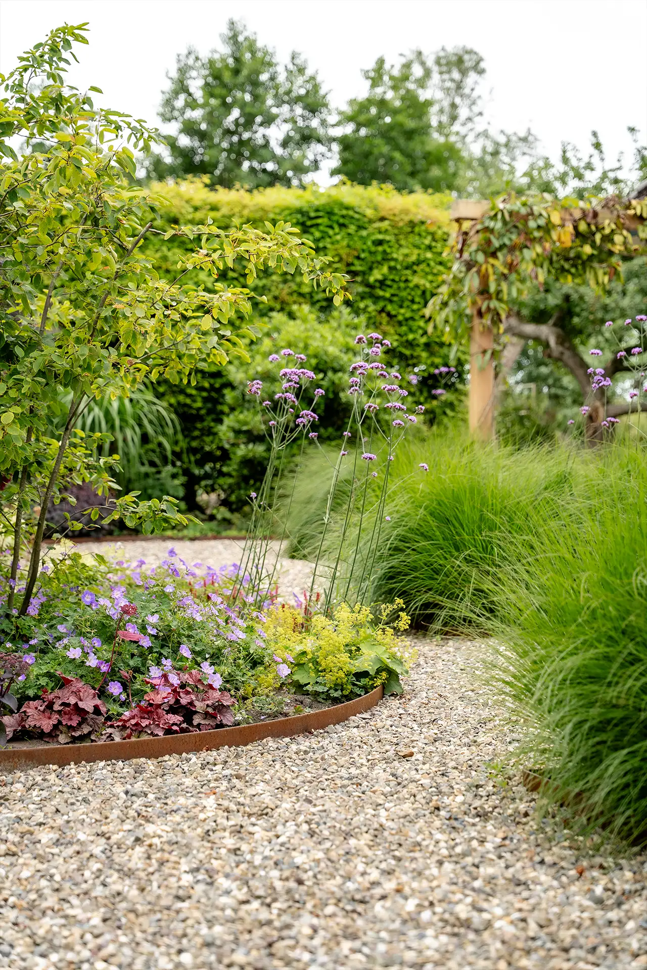 Sierlijke tuin met een gebogen grindpad, omgeven door kleurrijke bloemen, groene struiken en een pergola.