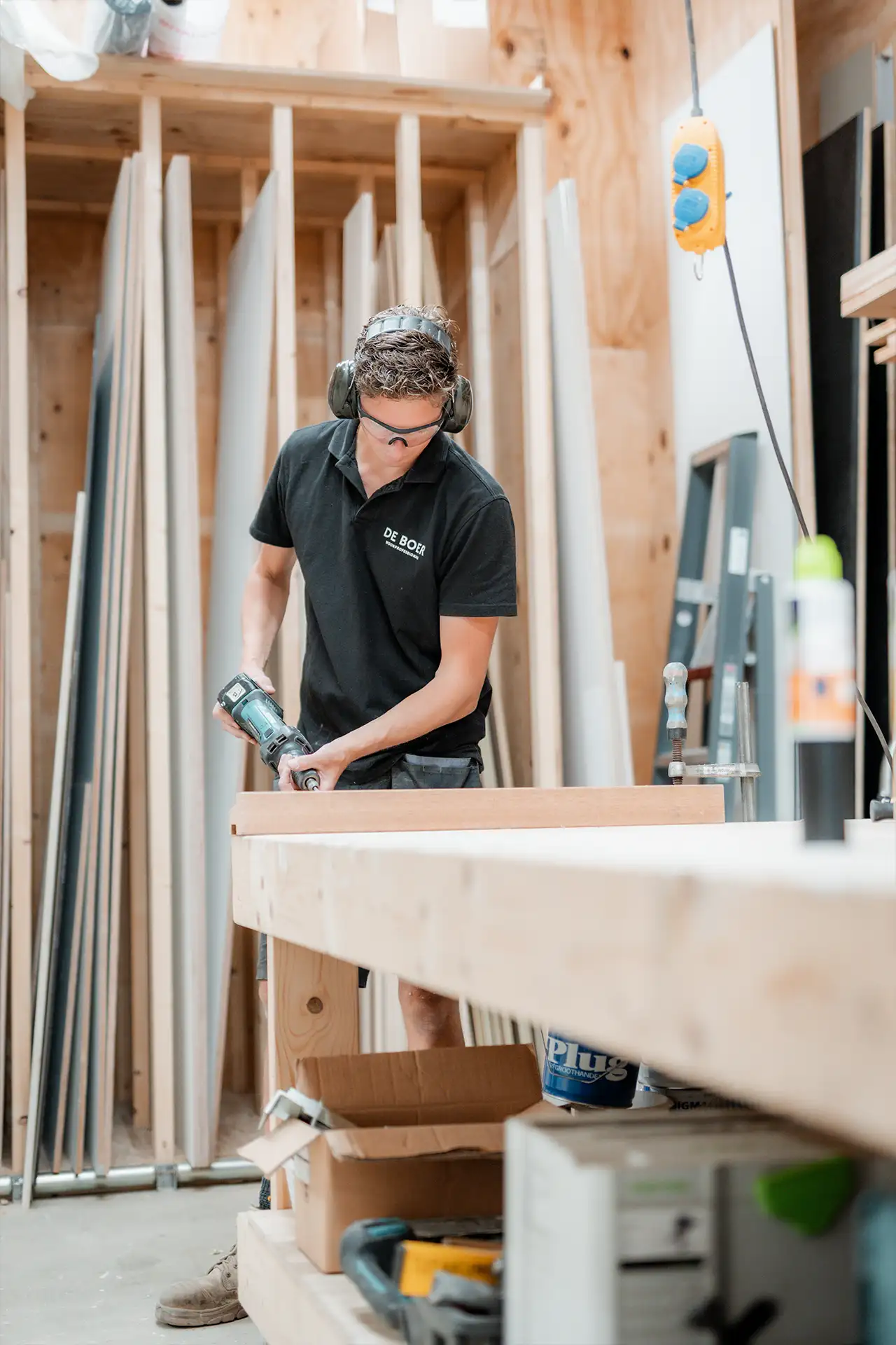 Man met veiligheidsbril en gehoorbescherming die werkt met een elektrische boor op een houten werkbank in een timmerwerkplaats.