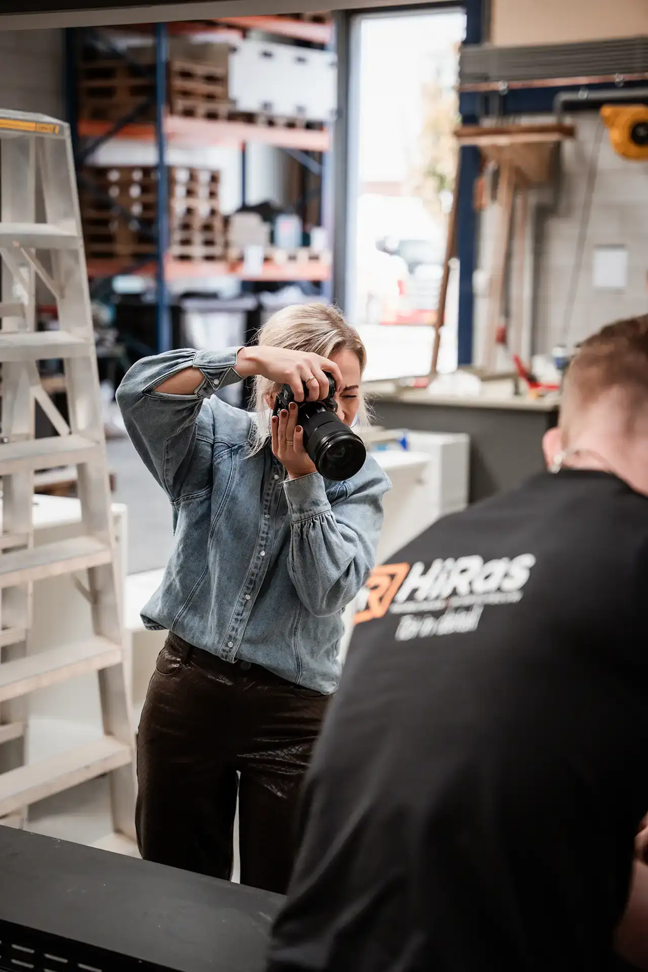 Vrouw met een spijkerblouse die binnenshuis een foto maakt met een camera gericht op een persoon met een zwart shirt met tekst.