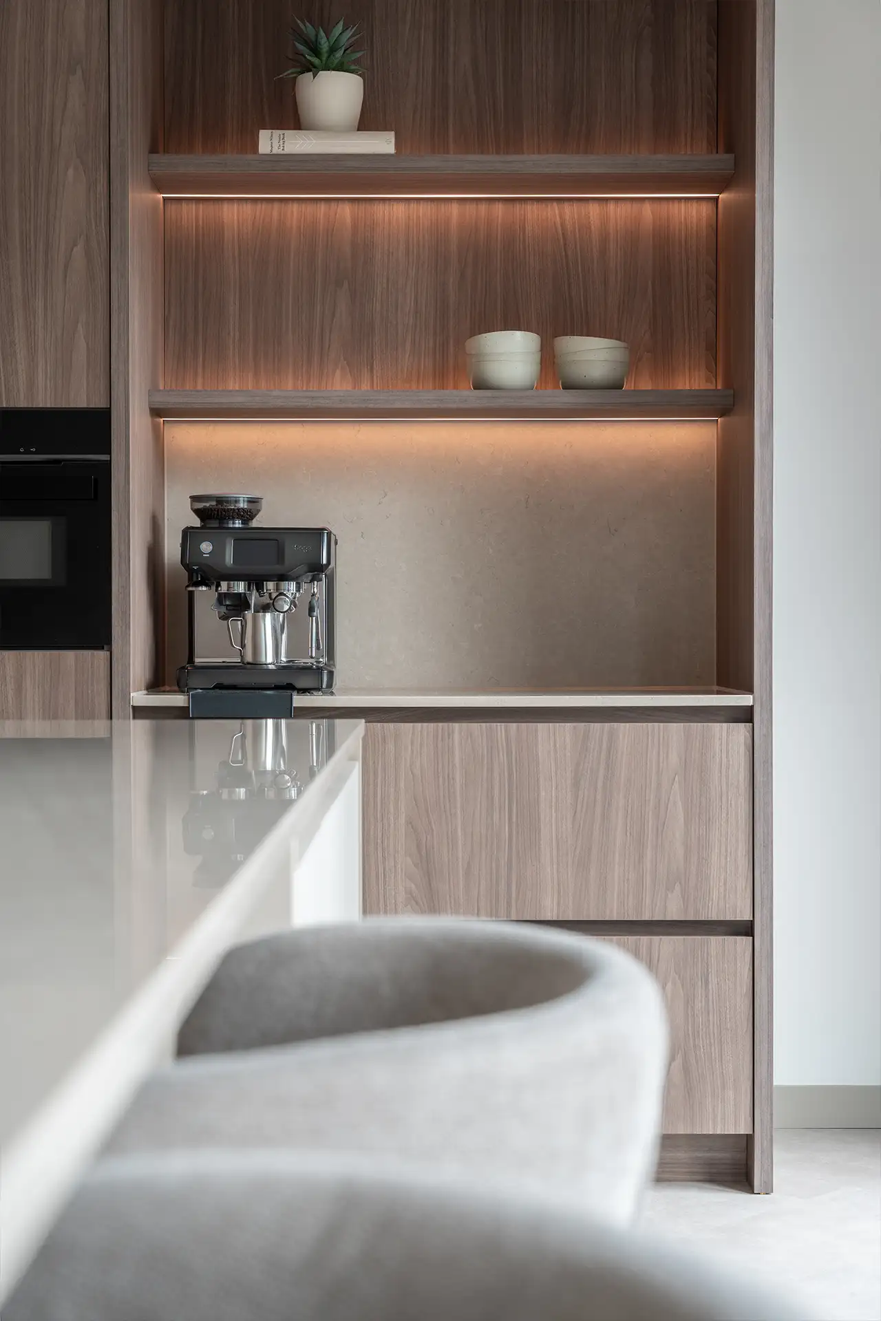 Moderne keuken met houten kast, ingebouwde koffiezetapparaat, twee keramische potten op een plank en een grijze stoel op de voorgrond.