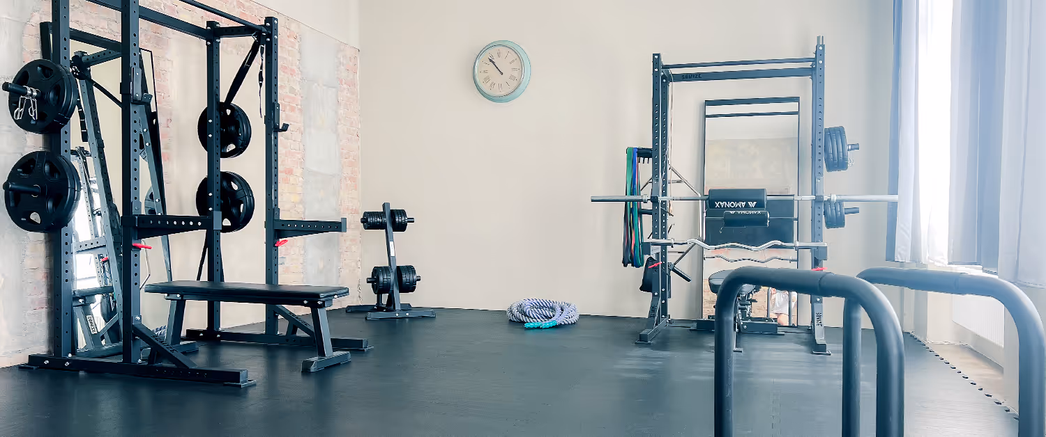 Helles Fitnessstudio mit Hantelbank, Power Rack, Gewichten und Trainingsseilen vor einer Wand mit Uhr