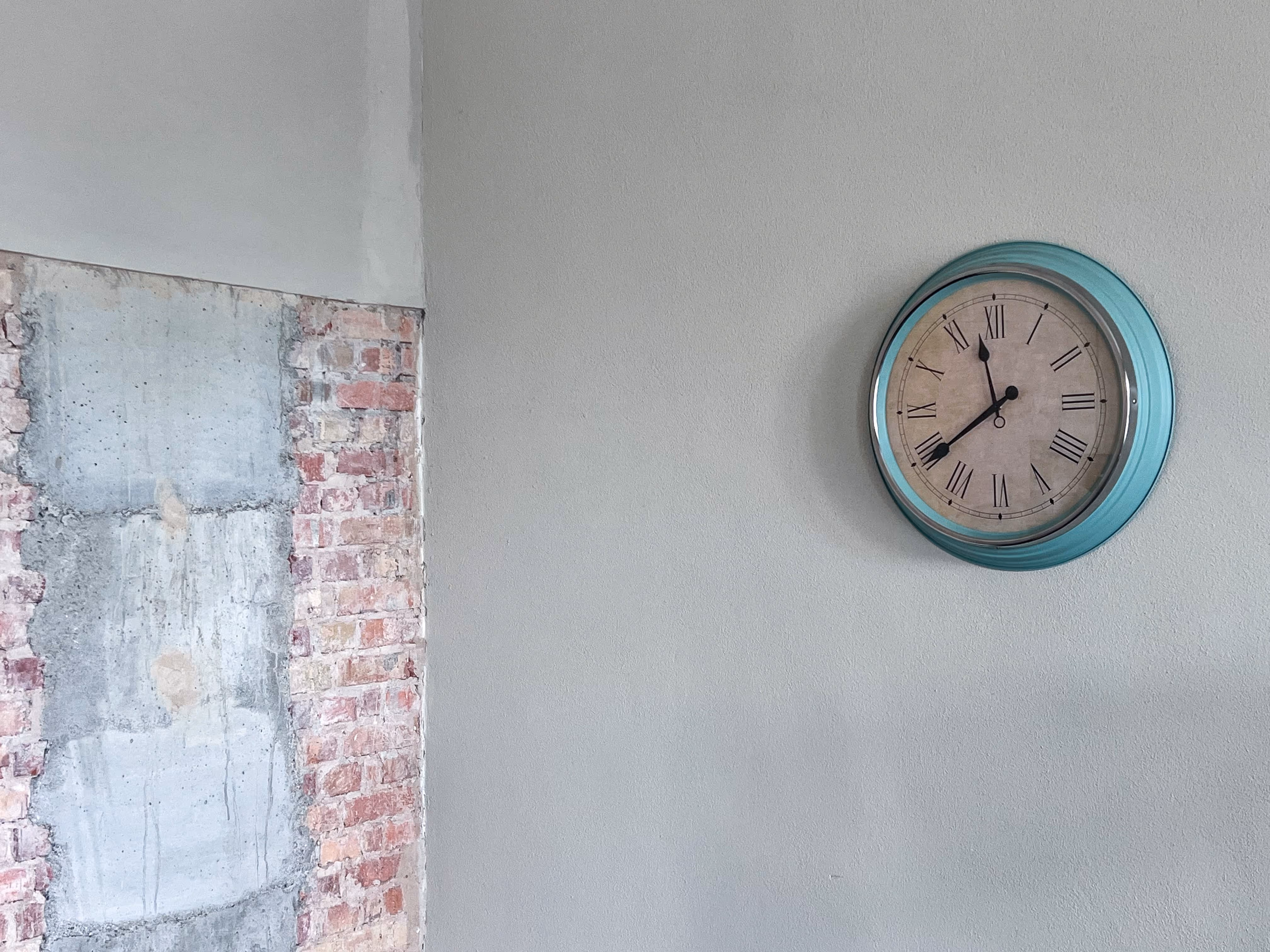 Runde Wanduhr mit türkisem Rahmen, an einer hellen Wand mit freigelegtem Mauerwerk