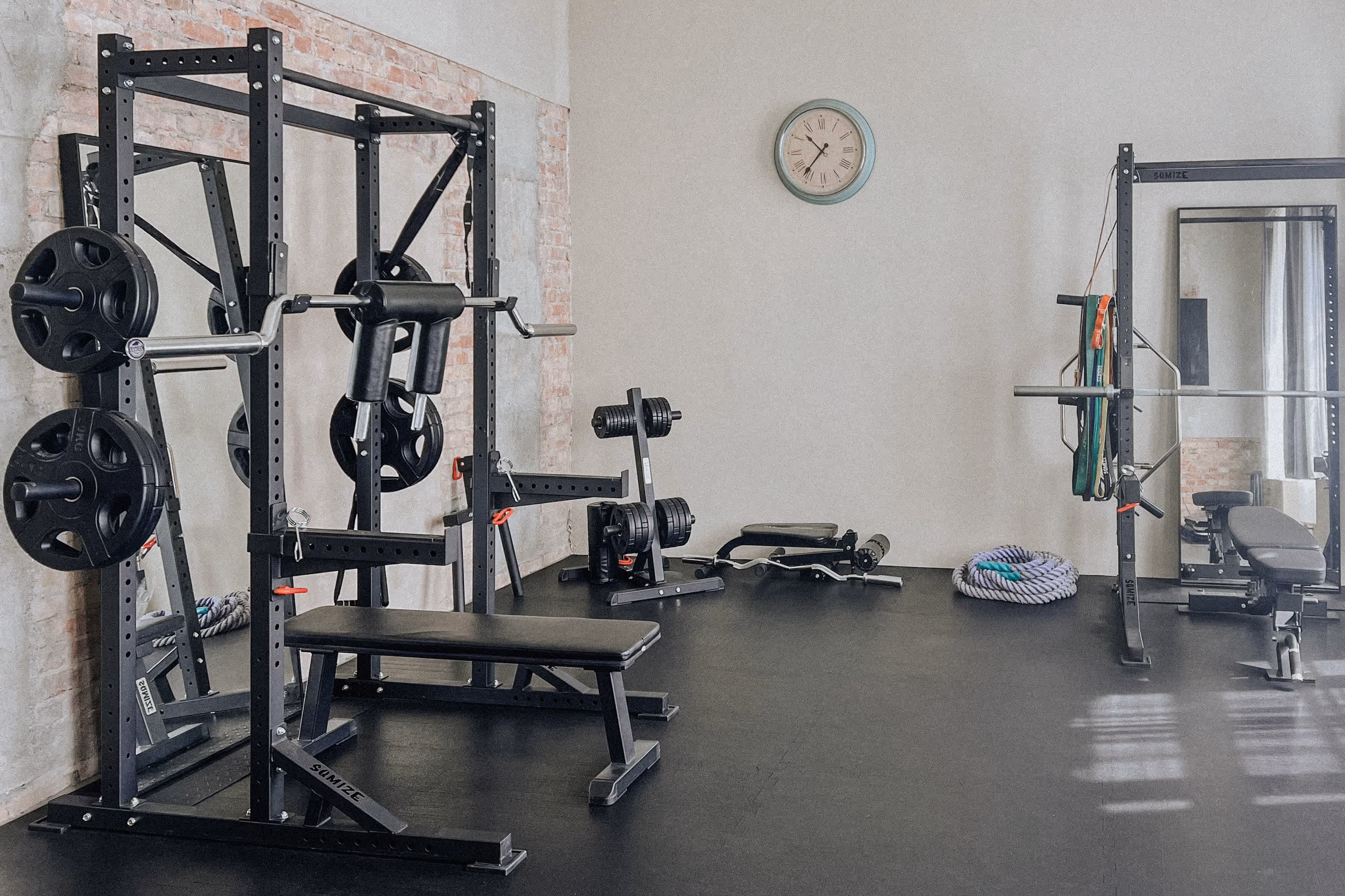 Fitnessraum mit Hantelbank, Power Racks, Gewichten, Trainingsseilen und Wanduhr vor einer Wand mit Sichtmauerwerk.