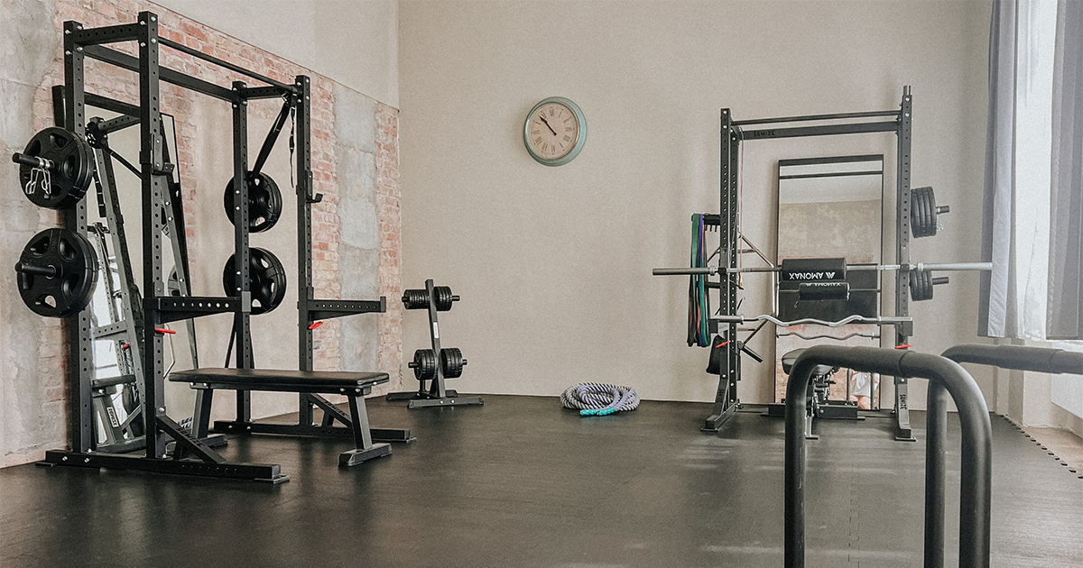 Hell beleuchteter Fitnessraum mit Hantelbänken, Gewichtsscheiben, Widerstandsbändern und einer Uhr an der Wand.