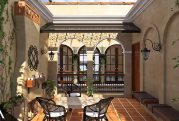 Sunlit Mediterranean-style exterior patio with terracotta tiles, two black wicker chairs, wall-mounted lanterns, and archway overlooking a balcony with greenery.