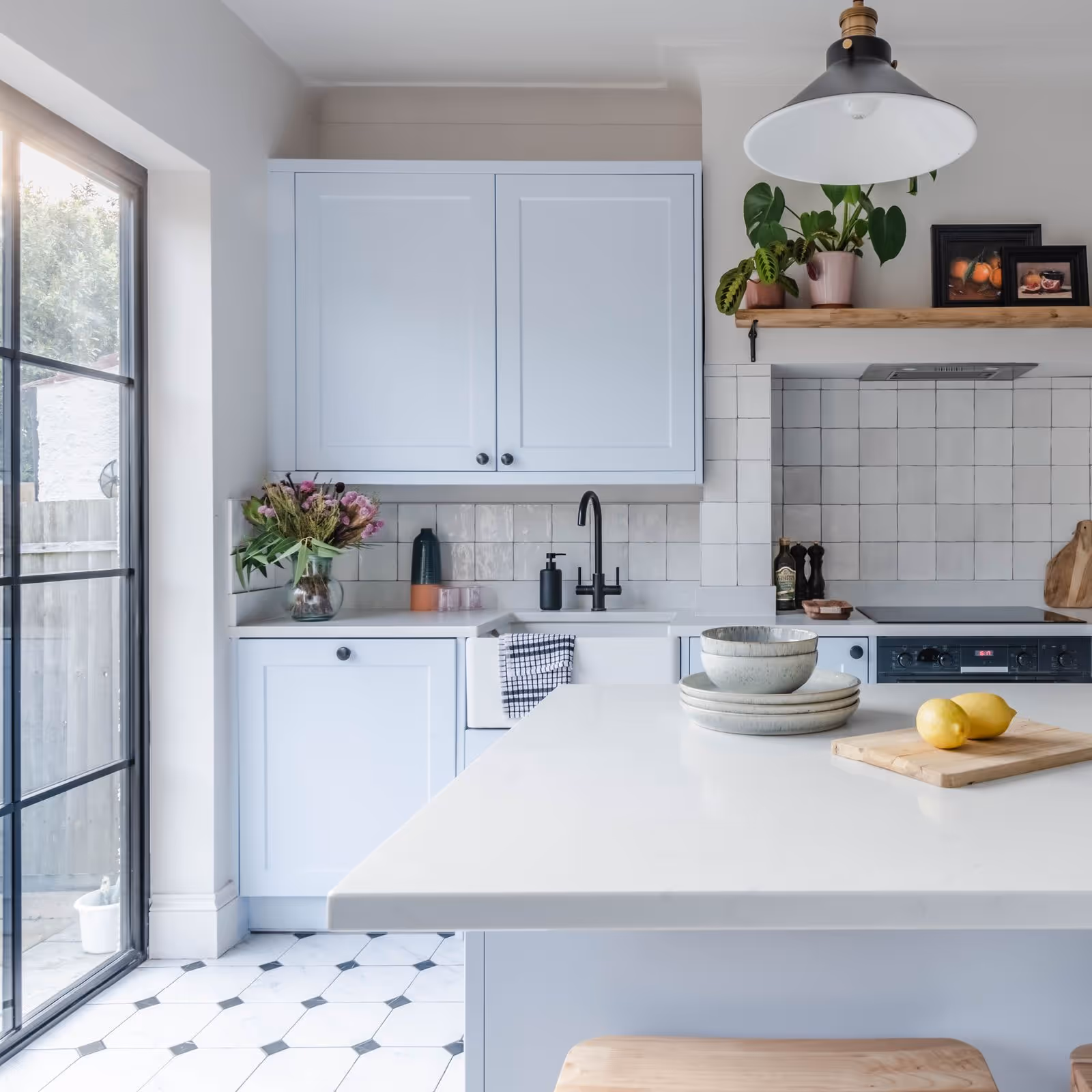 Bright modern kitchen with light blue cabinets, white tiled backsplash, island with dishes and lemons, and large window.