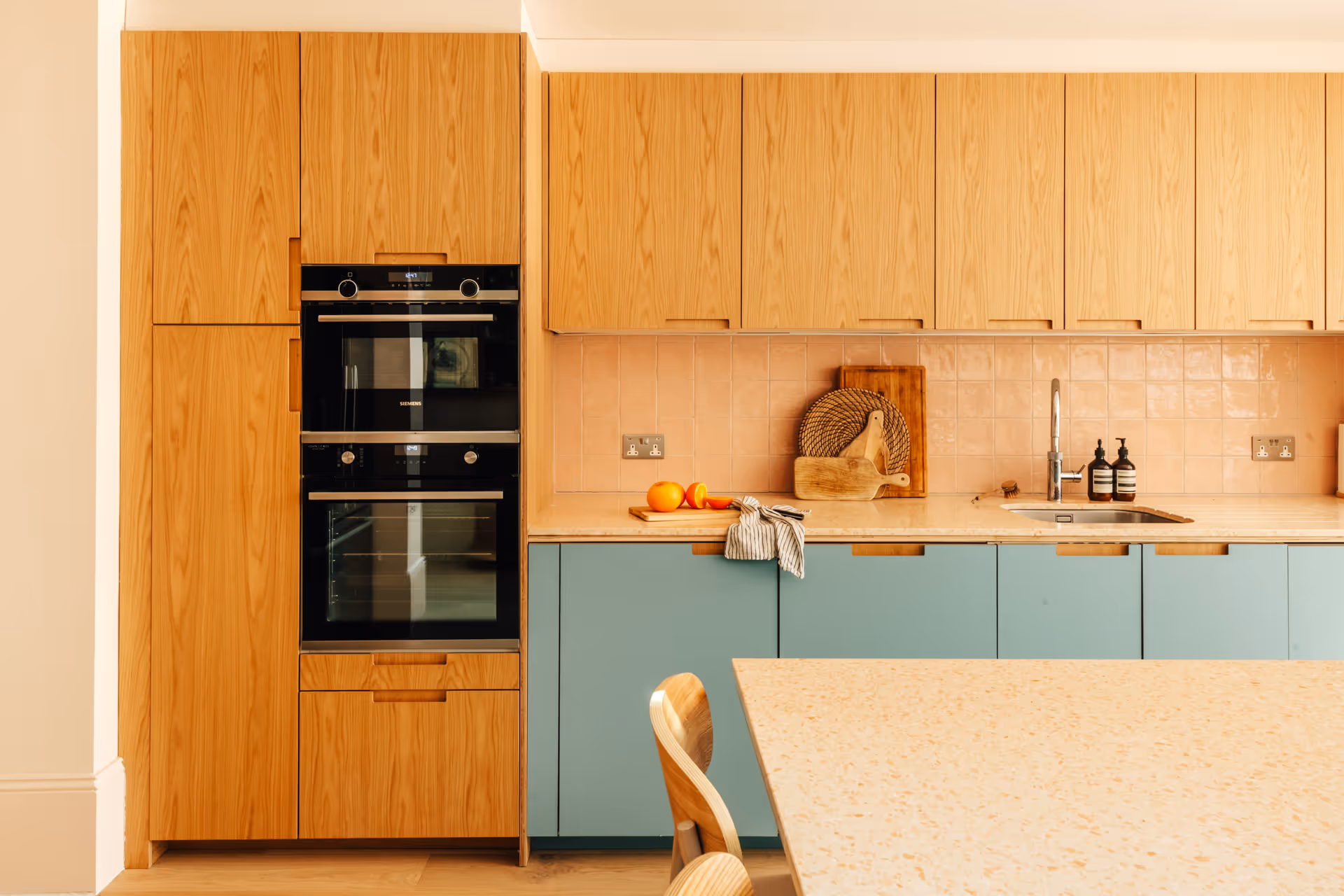Modern kitchen with light wood upper cabinets, teal lower cabinets, built-in double oven, and a beige countertop.