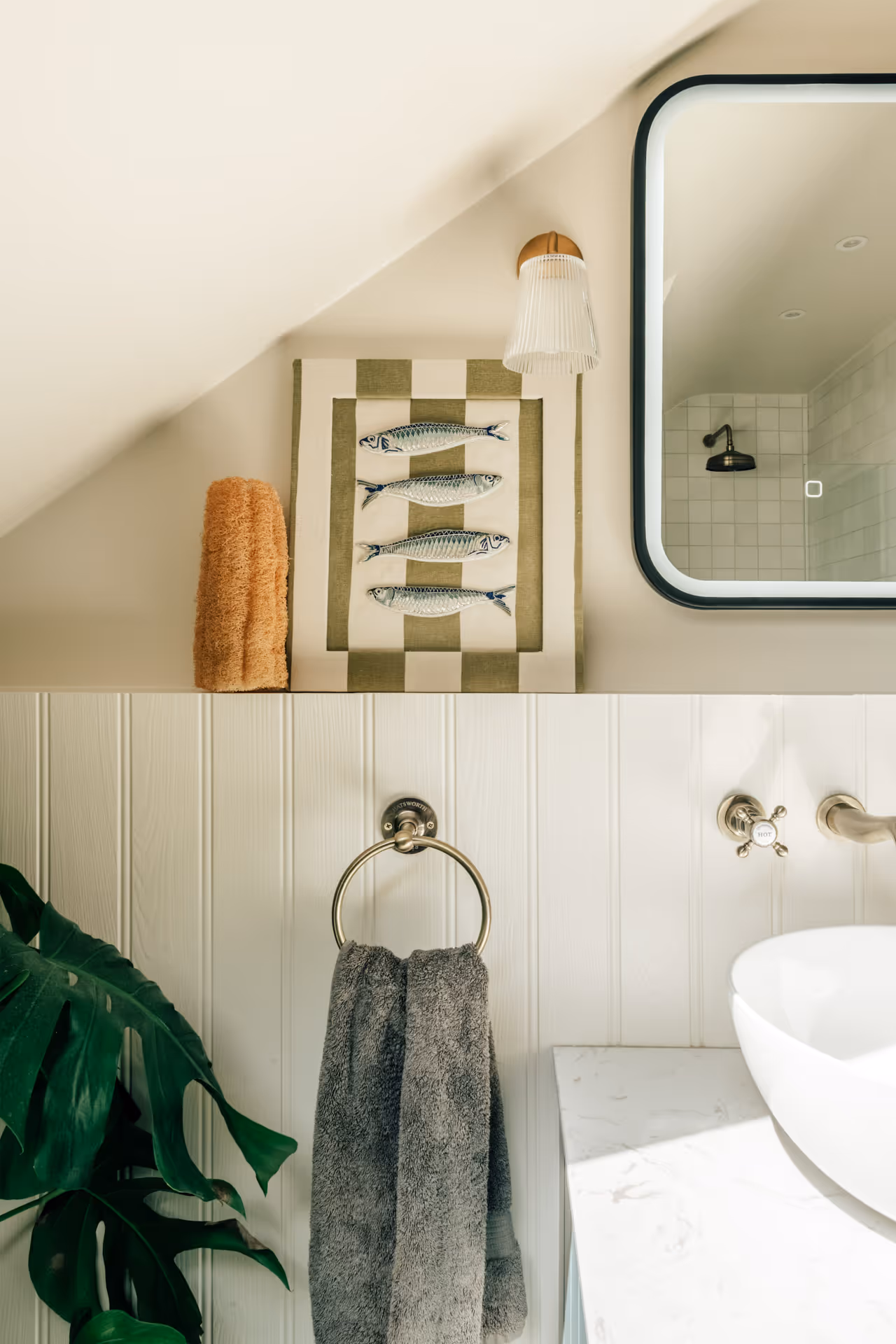 Bathroom interior showing a towel ring with a gray towel, a framed art piece of four fish, a tan rolled towel, a large green plant, and a square mirror reflecting a showerhead.