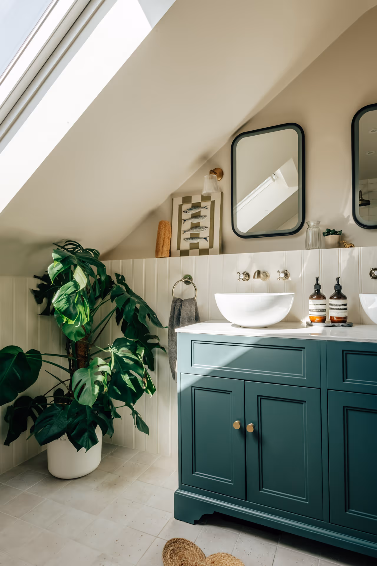 Bathroom vanity with teal cabinets, white vessel sink, dual mirrors, and a large potted green plant under a skylight.