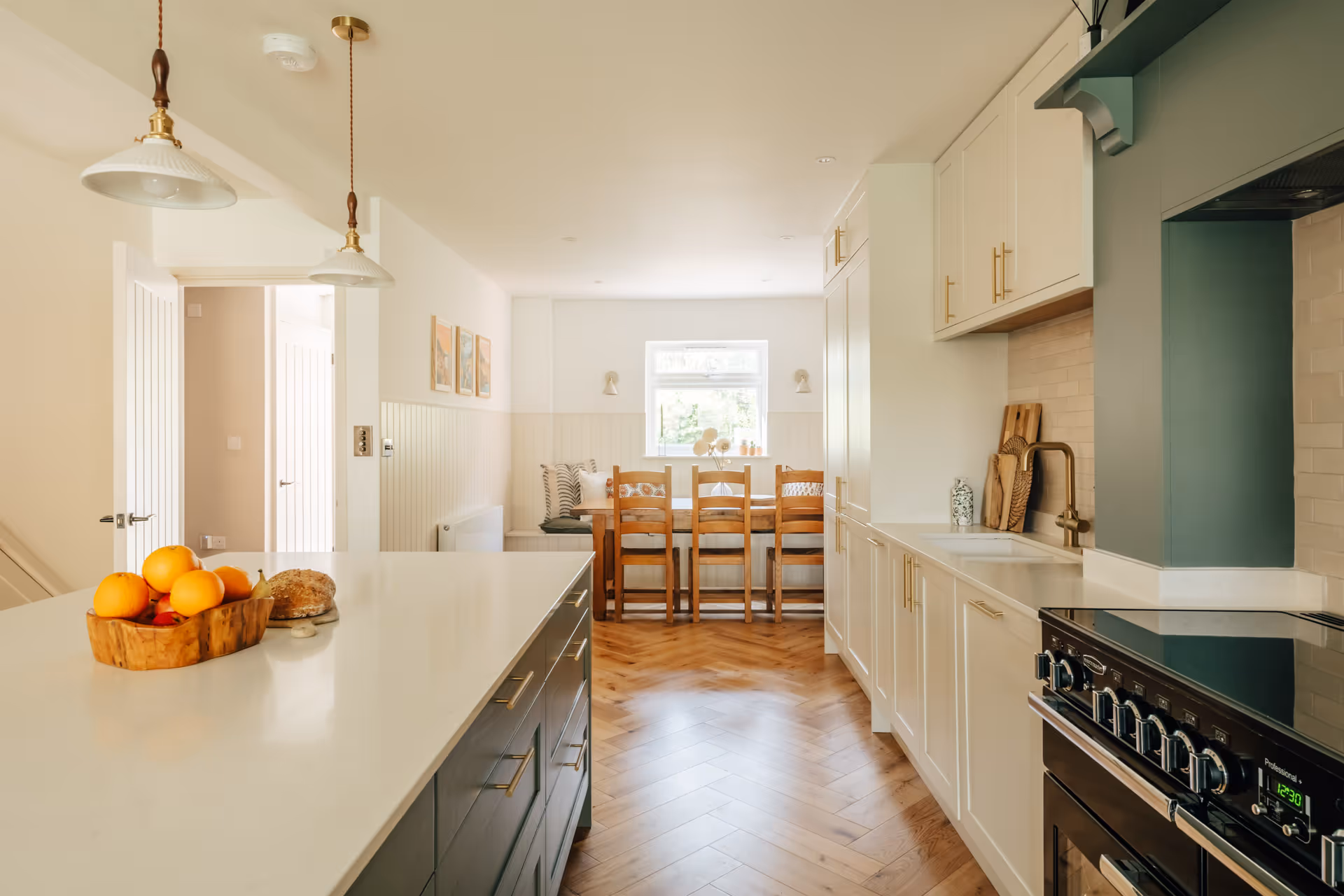 Modern kitchen with white countertops, dark island holding fruit bowl, wooden dining table with chairs by window, and herringbone wood floor.