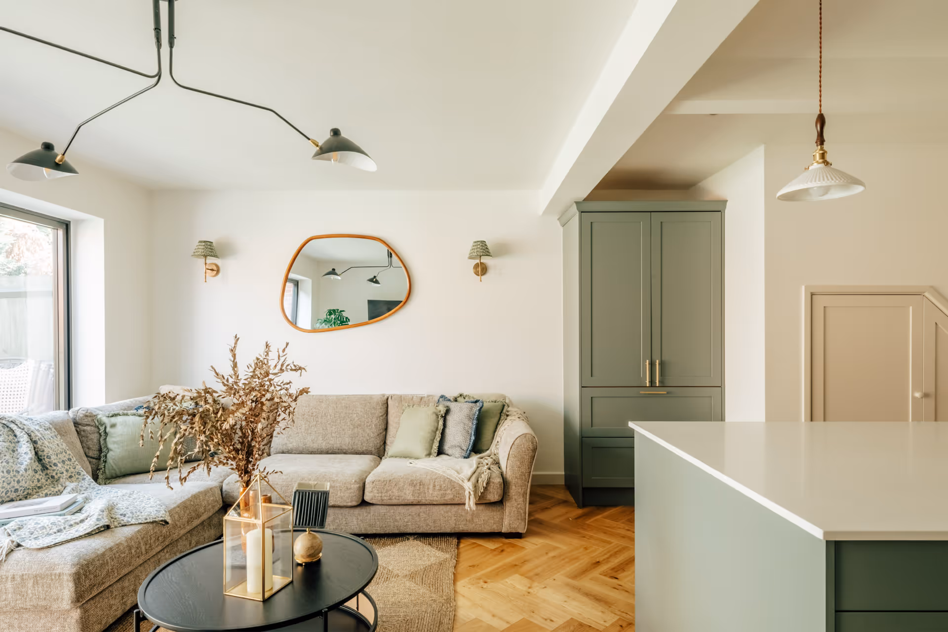 Cozy living room with beige sectional sofa, decorative pillows, round black coffee table with dried flowers and candles, green cabinet, and light wood flooring.