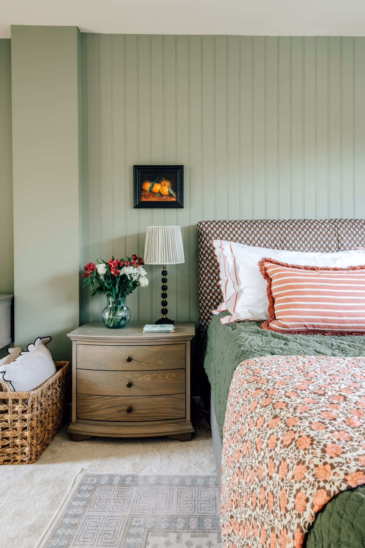 Bedroom corner with green paneled wall, wooden nightstand holding a lamp and vase of red and white flowers, patterned bed with striped and ruffled pillows, and a woven basket with white cushions on beige carpet.