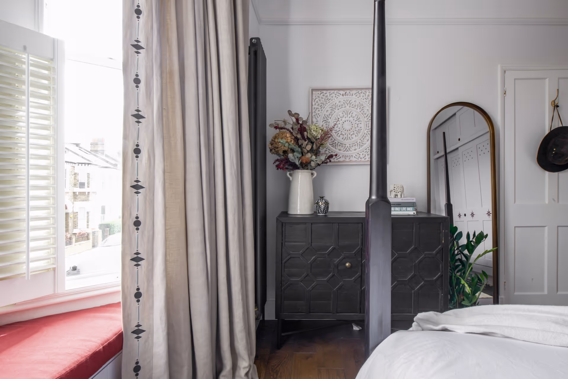 Bedroom corner with a dark wooden cabinet topped with a white vase of dried flowers, decorative wall art, and an arched mirror reflecting a closet.