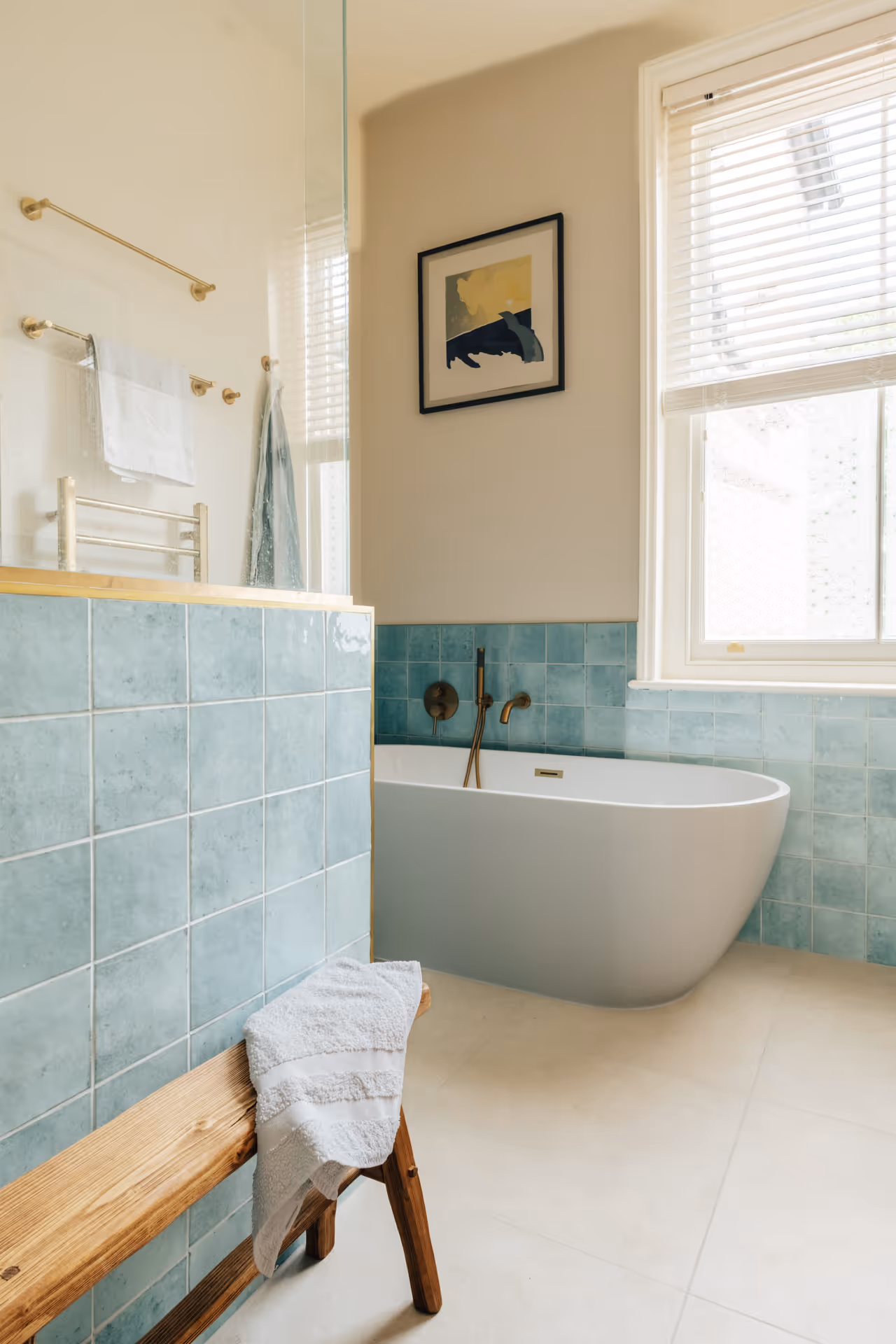 Modern bathroom featuring a freestanding white bathtub with brass fixtures, blue tiled walls, a wooden bench with a white towel, and a window with white blinds.
