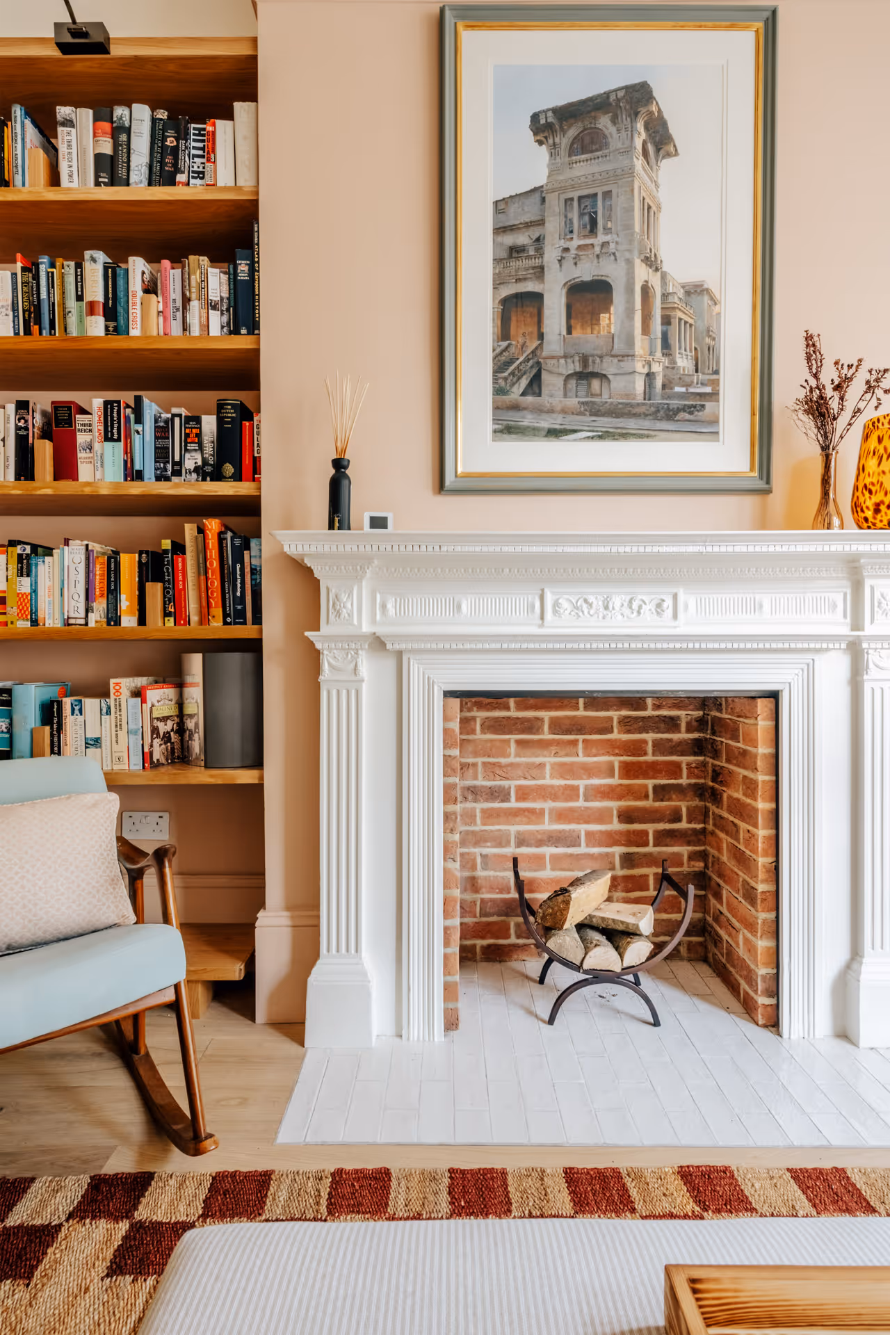 Cozy living room corner with a white ornate fireplace containing stacked firewood, a light blue rocking chair, wooden bookshelf, and framed architectural print above the mantel.