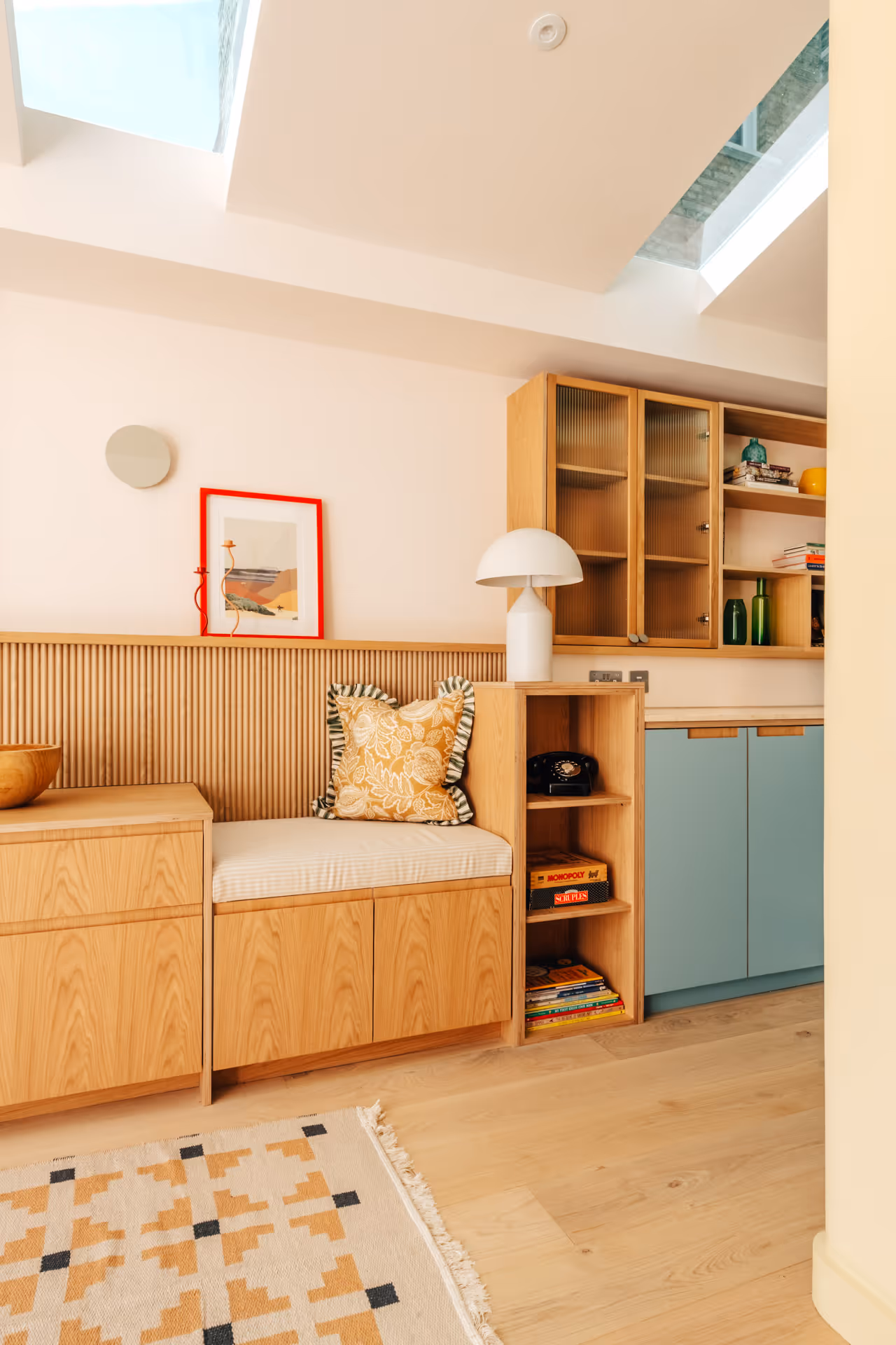 Cozy corner with wooden built-in bench with a floral cushion, lamp, vintage phone, and shelves with books and decor in a bright room with skylights.
