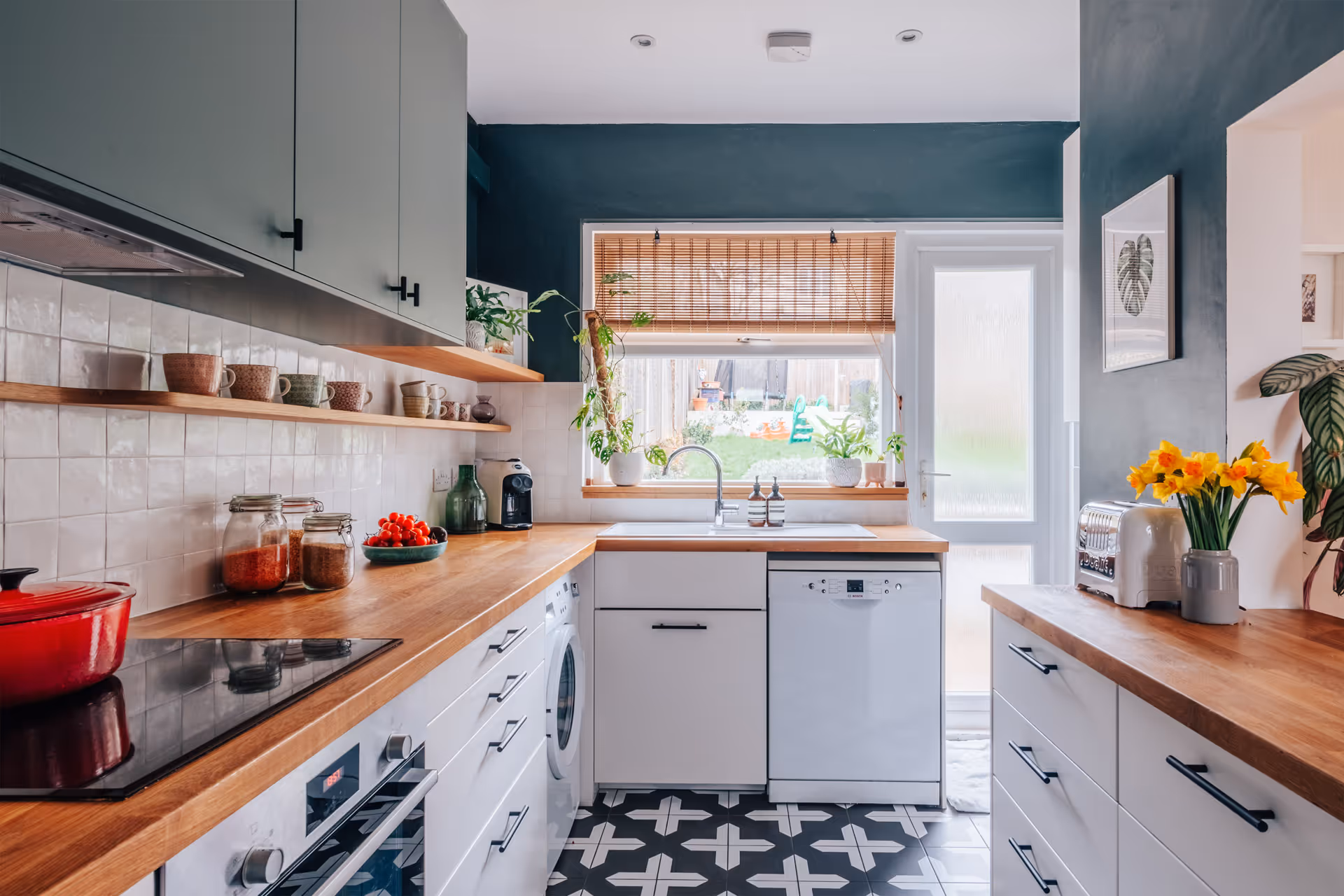 Modern kitchen with wooden countertops, white cabinets, patterned black-and-white floor tiles, and a window with bamboo blinds overlooking a garden.