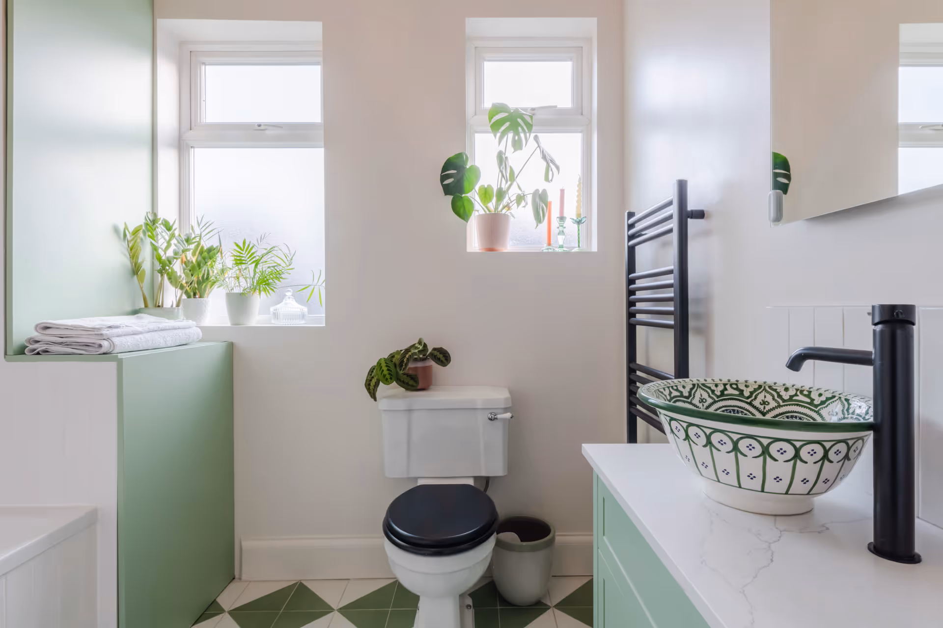 Bright bathroom with white walls, green tiled floor, white toilet with black seat, patterned round sink, and several potted plants on windowsills and countertop.