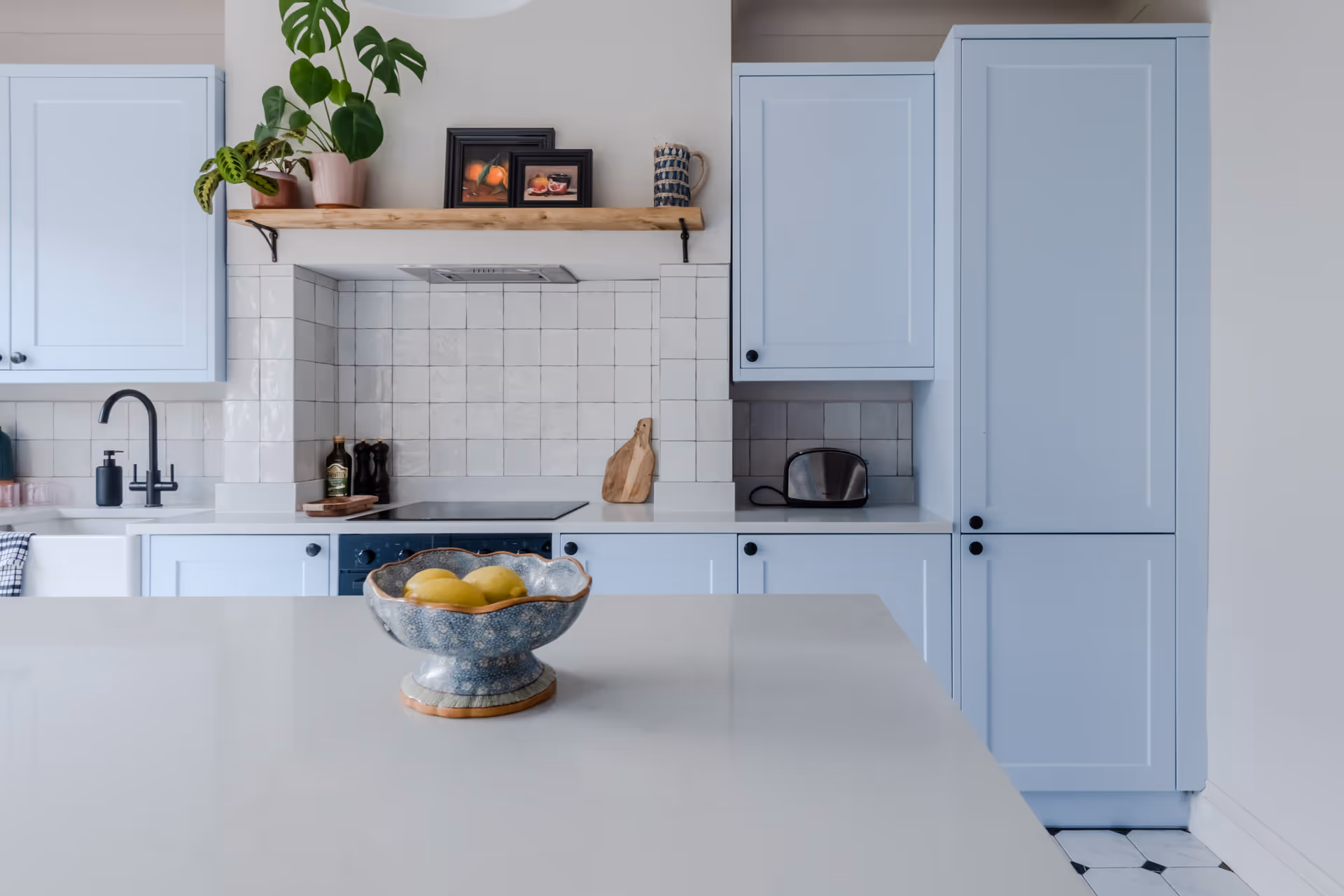 Modern kitchen with light blue cabinets, white tiled backsplash, black faucet, stovetop, and a blue fruit bowl with yellow lemons on a white island.