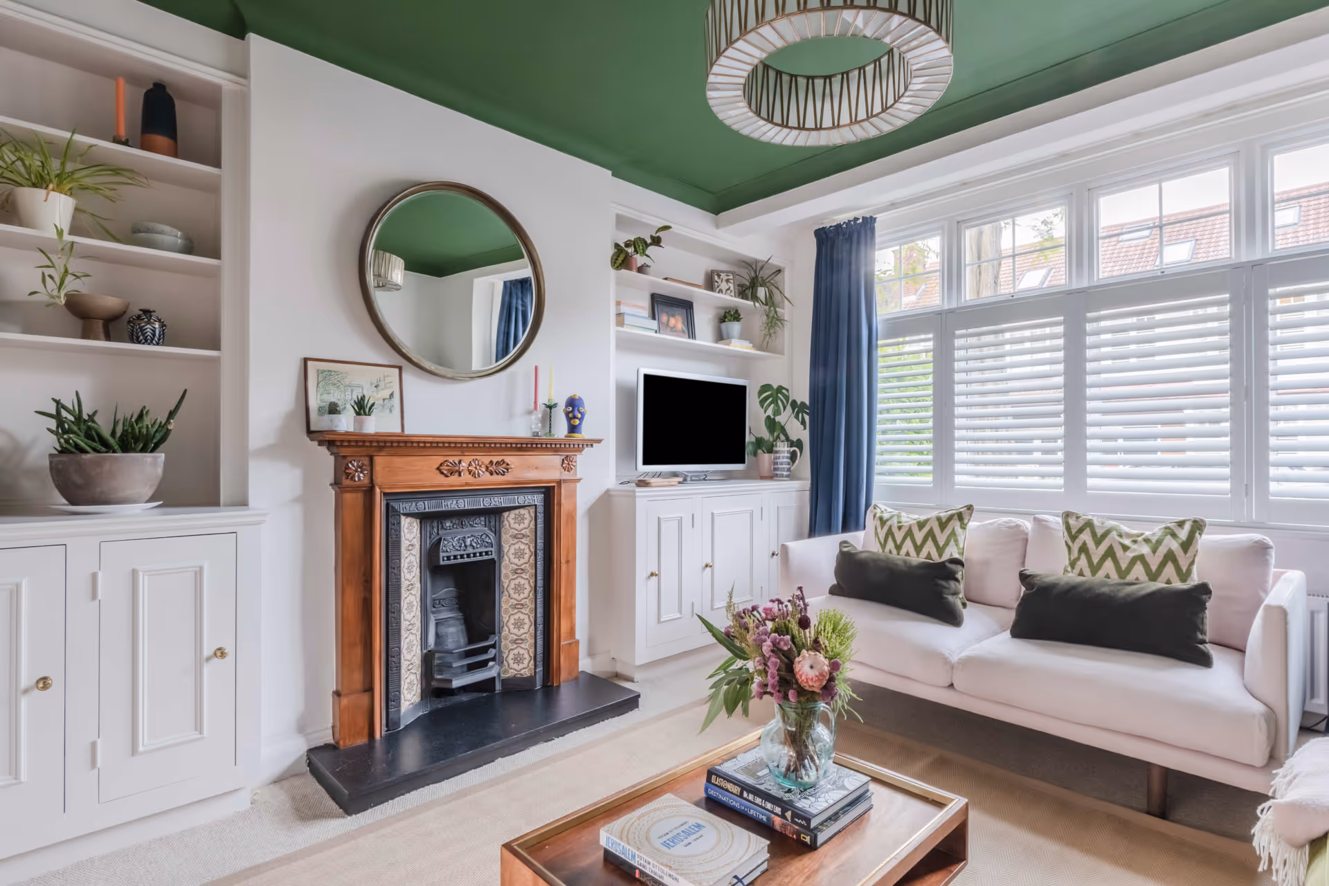 Bright living room with a white sofa with green and beige cushions, a wooden fireplace with a round mirror above, built-in shelves, and large windows with white shutters and blue curtains.