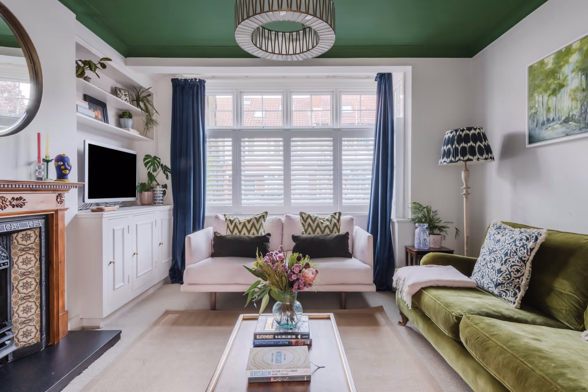 Living room with a green velvet sofa, white loveseat, bay window with blue curtains, patterned cushions, and a glass-top coffee table with a flower vase.