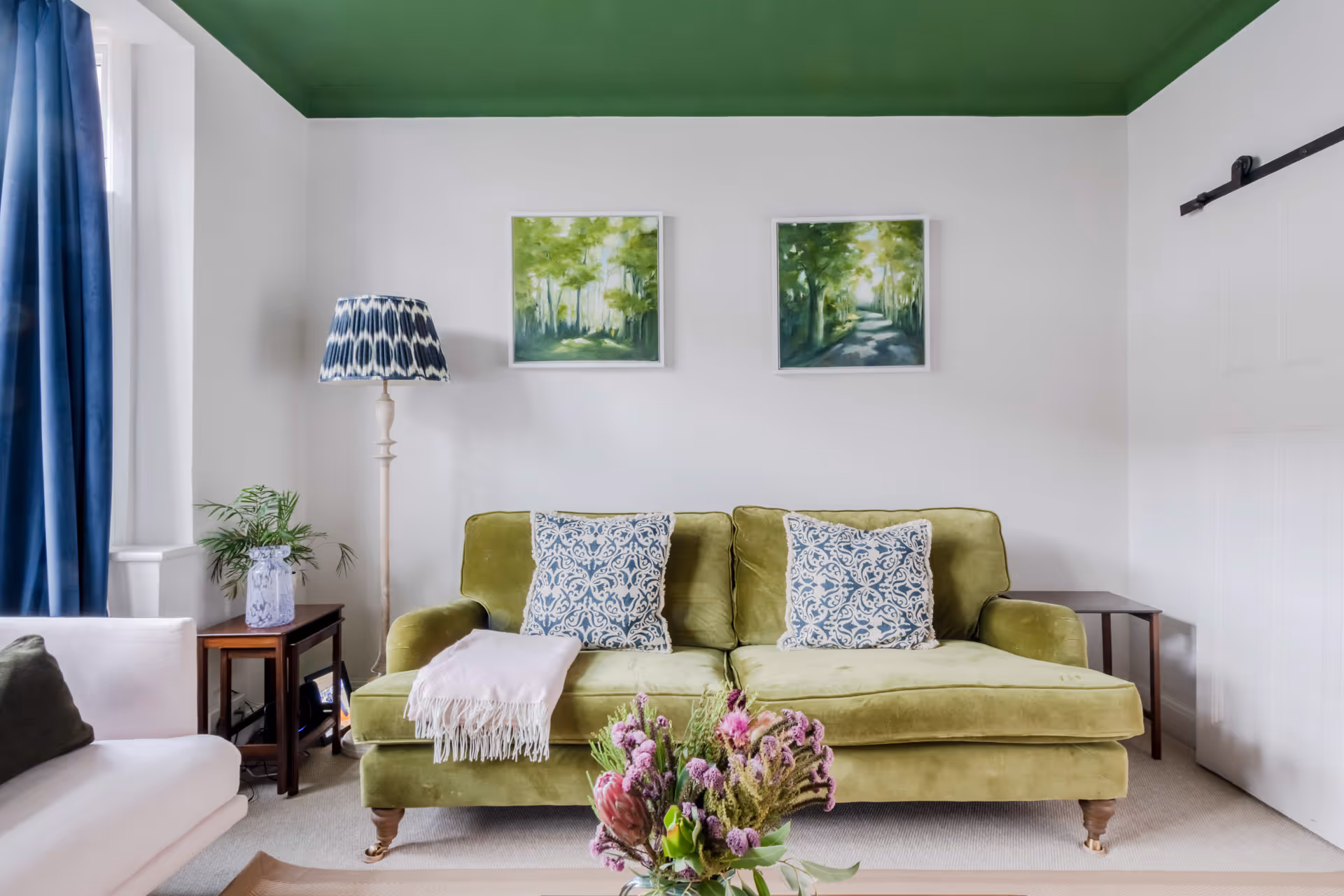 Living room with a green velvet sofa, patterned blue and white pillows, a white throw, floor lamp with blue patterned shade, two green-themed paintings, and a bouquet of flowers on a coffee table.
