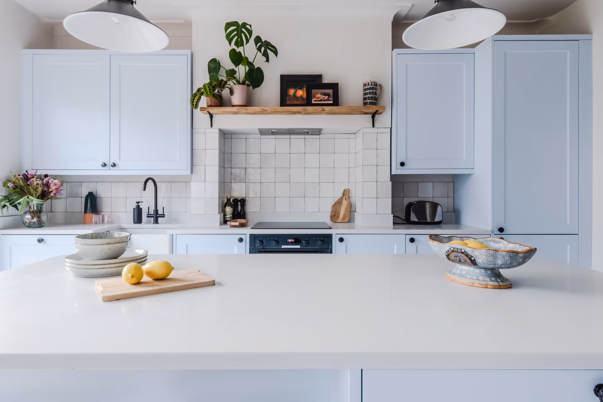 Modern kitchen with light blue cabinets, a white countertop island with lemons, stacked plates, and a decorative bowl, and a wooden shelf holding plants and framed pictures.