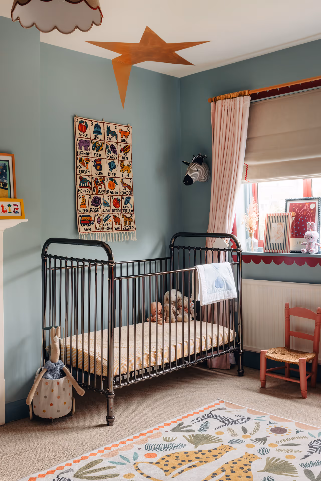 Children's bedroom with black metal crib, stuffed animals inside, pastel blue walls, educational wall hanging, and colorful animal rug.