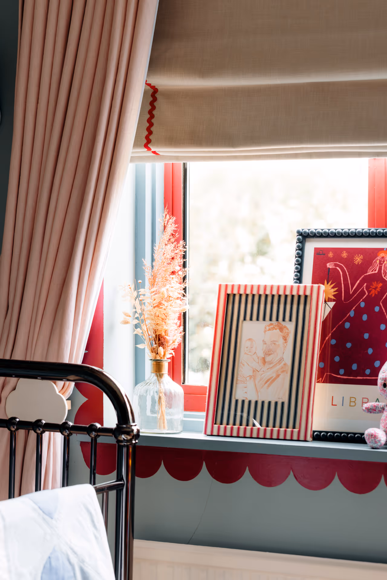 Window with beige roman shade partially lowered, pink curtains, dried flowers in a glass vase, and framed pictures on the windowsill next to a black metal bedframe.